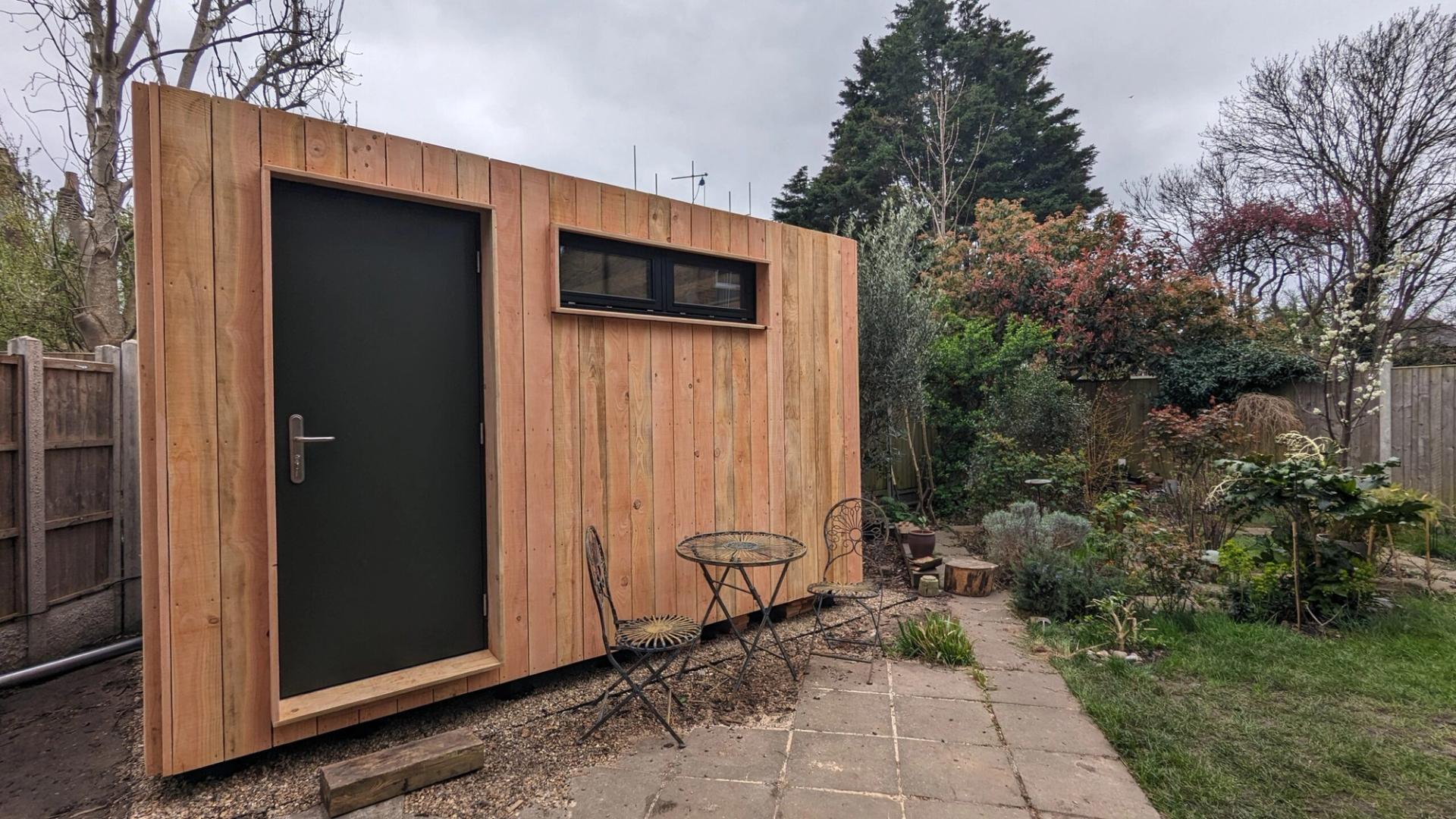 A small modular garden shed built from spruce plywood with a dark door and narrow window stands beside a metal table and two chairs in a fenced, landscaped back garden with trees and shrubs.