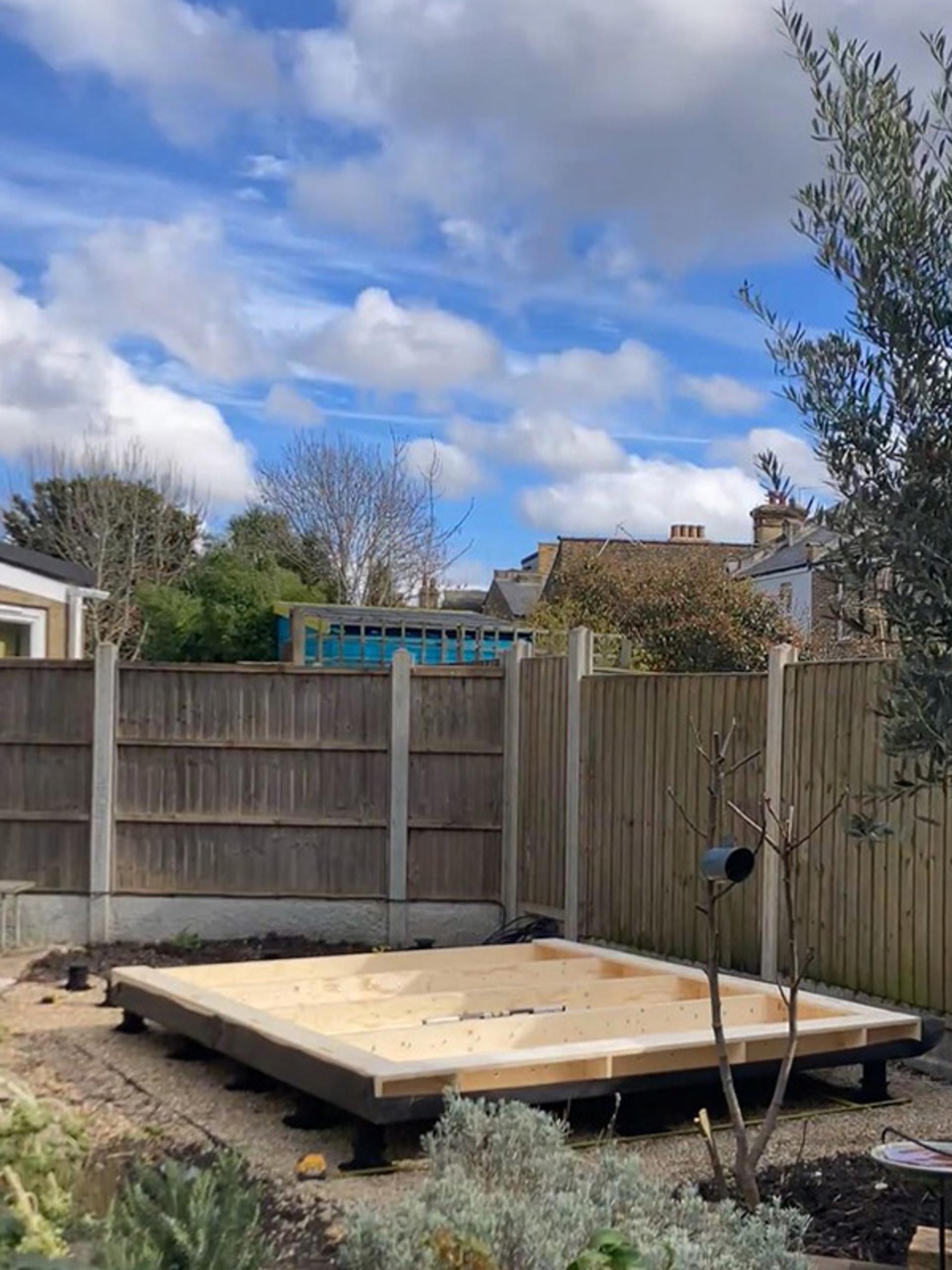 A sustainable wooden raised garden bed frame, possibly crafted from birch plywood, sits on gravel in a back garden, surrounded by fencing and greenery, under a blue sky with scattered clouds.