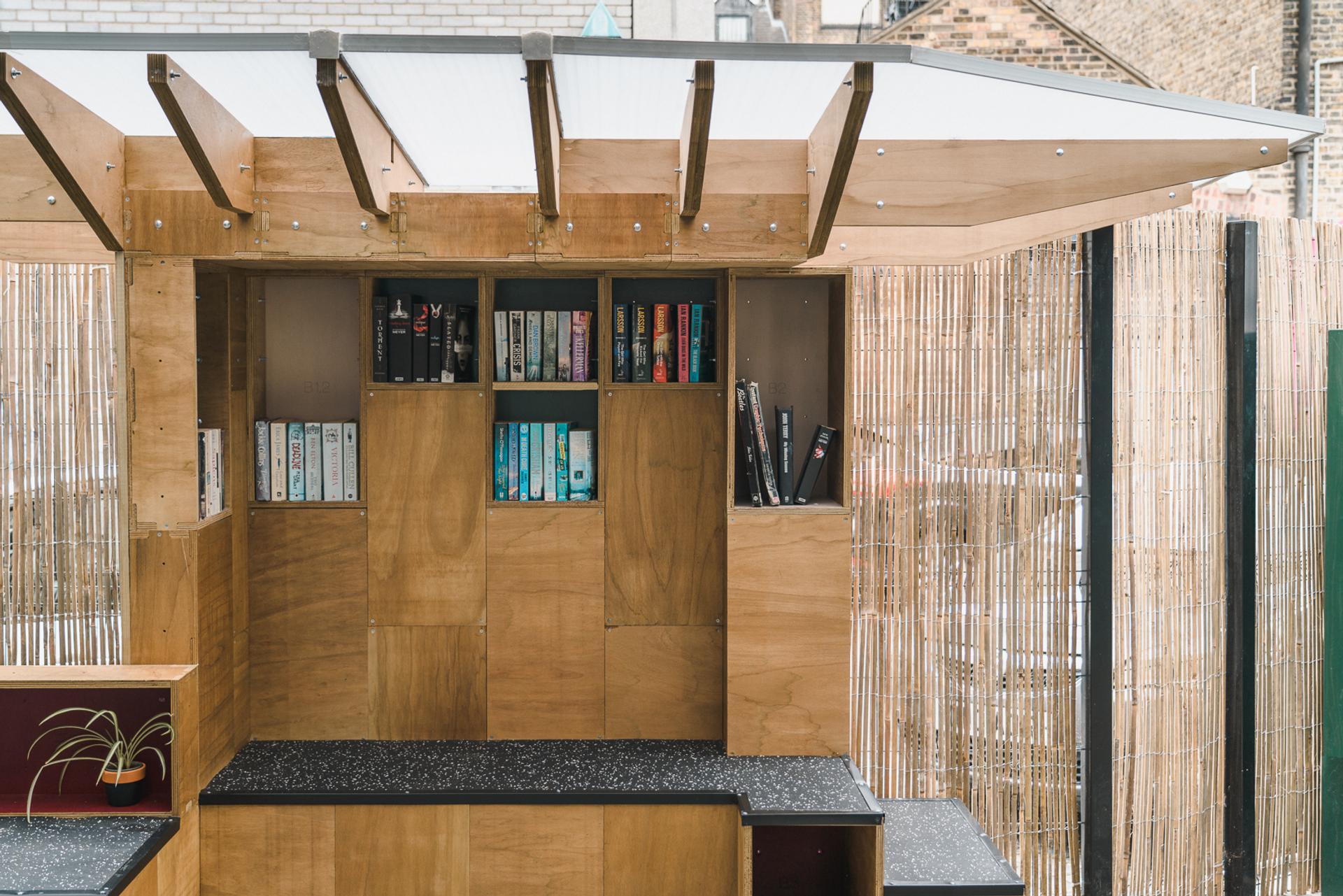 A small outdoor wooden library with shelves holding a variety of books, partially enclosed by bamboo fencing and a translucent roof. Built-in benches and the black worktop highlight the space’s recycle and upcycle ethos.