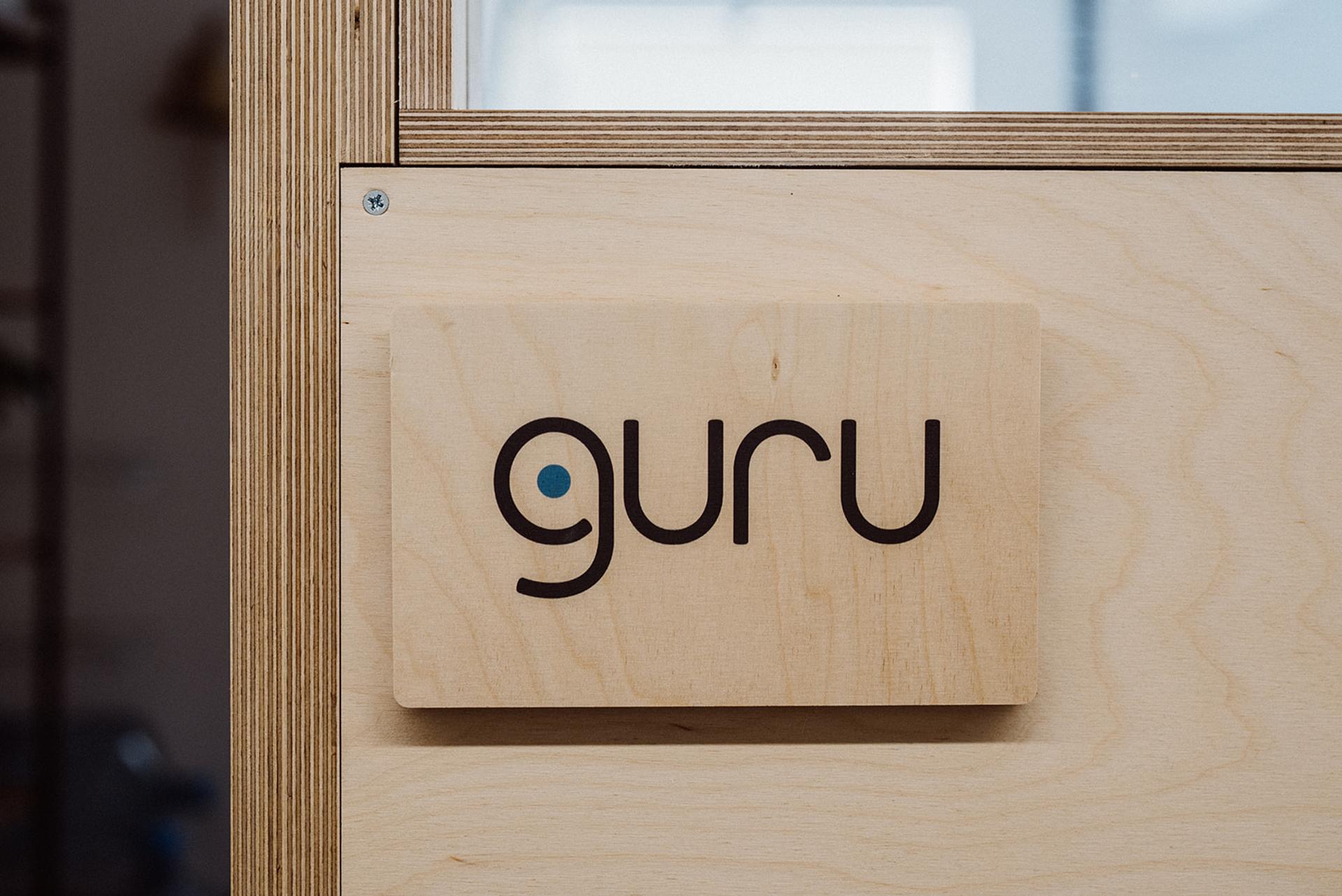 A wooden sign made from birch with the word guru in lowercase black letters, where the letter g has a blue dot inside it, is mounted on a light wood wall with metal screws visible.