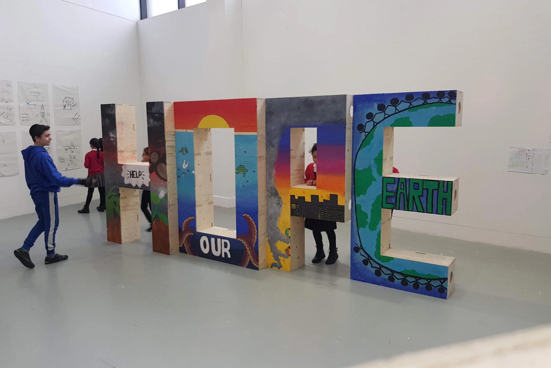 Children walk around and through large painted 3D letters spelling HOPE OUR EARTH in an art gallery, with colourful artwork on the letters inspired by upcycle themes and the circular economy to promote environmental awareness.
