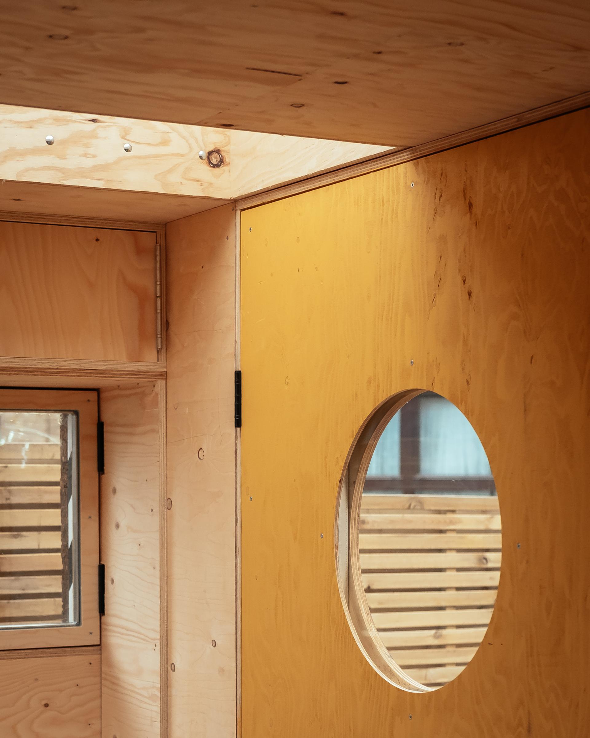 Interior view of a wooden structure with light plywood walls and ceiling showcases sustainability. A door with a round window and a small rectangular window are visible, whilst upcycled wooden slats outside offer privacy and allow natural light to enter.
