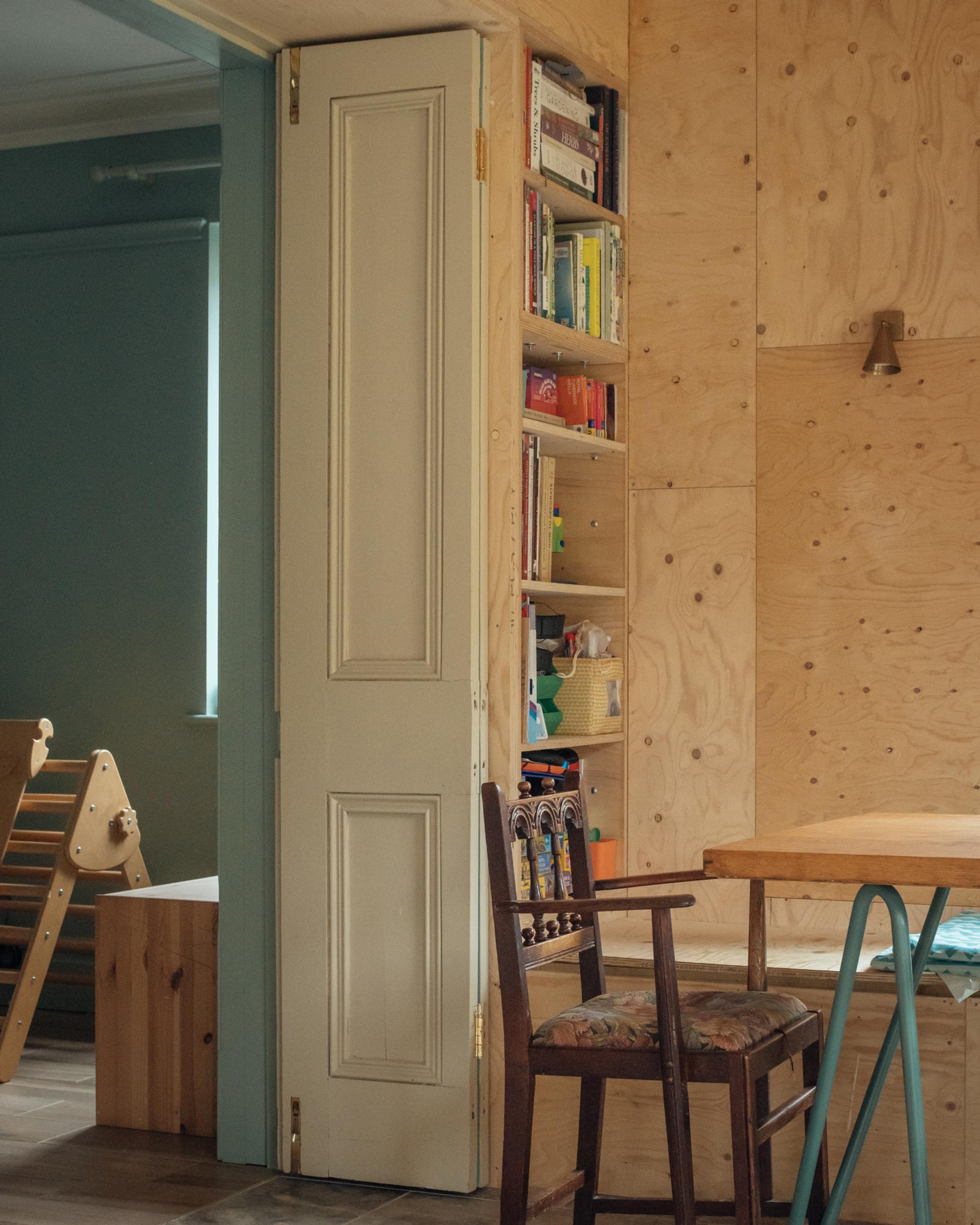A wooden dining chair sits next to a built-in bookcase integrated into a plywood wall constructed with a timber U-Build system.