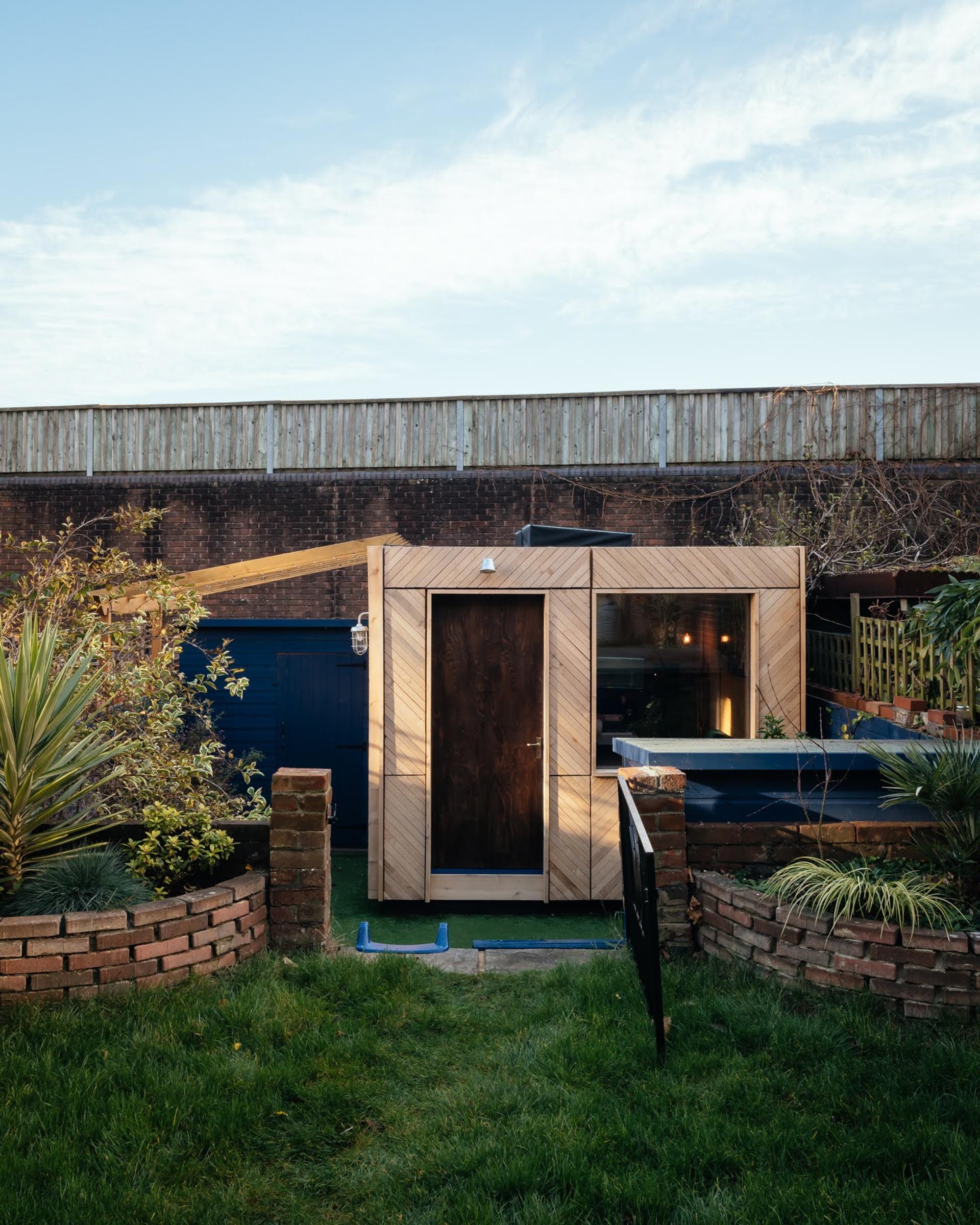 A small, sustainable wooden garden shed with a dark door and large window sits in a fenced back garden, surrounded by greenery and brick borders. A railway fence and blue sky are visible in the background.