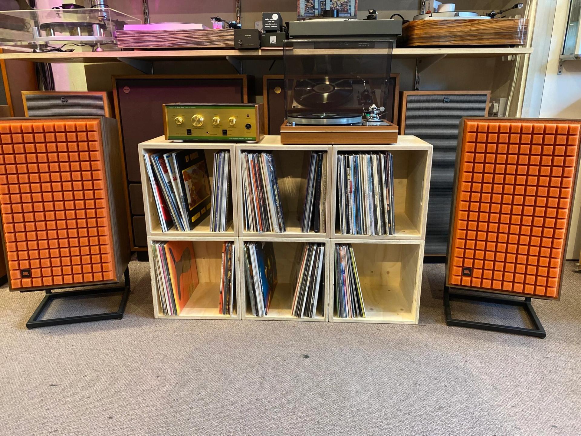 A turntable and vintage amplifier, both upcycled treasures, sit atop a wooden shelf filled with vinyl records. Two orange-speakered stereo speakers stand on either side, all set on a carpeted floor in a cosy room.