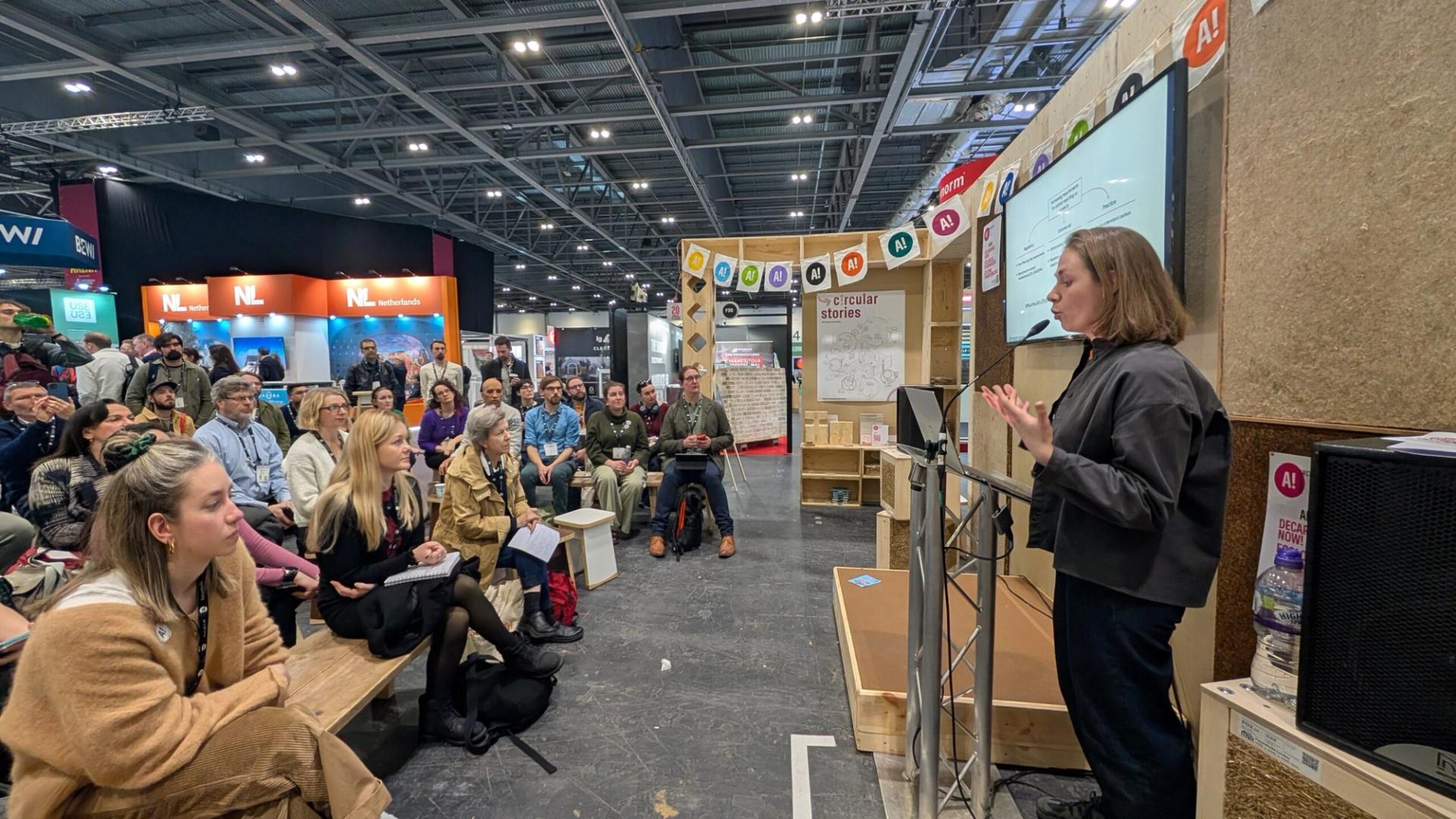 A woman gives a presentation to a seated audience at an indoor event focused on modular and timber construction. Display stands and banners are visible in the background, whilst people listen attentively, some taking notes.