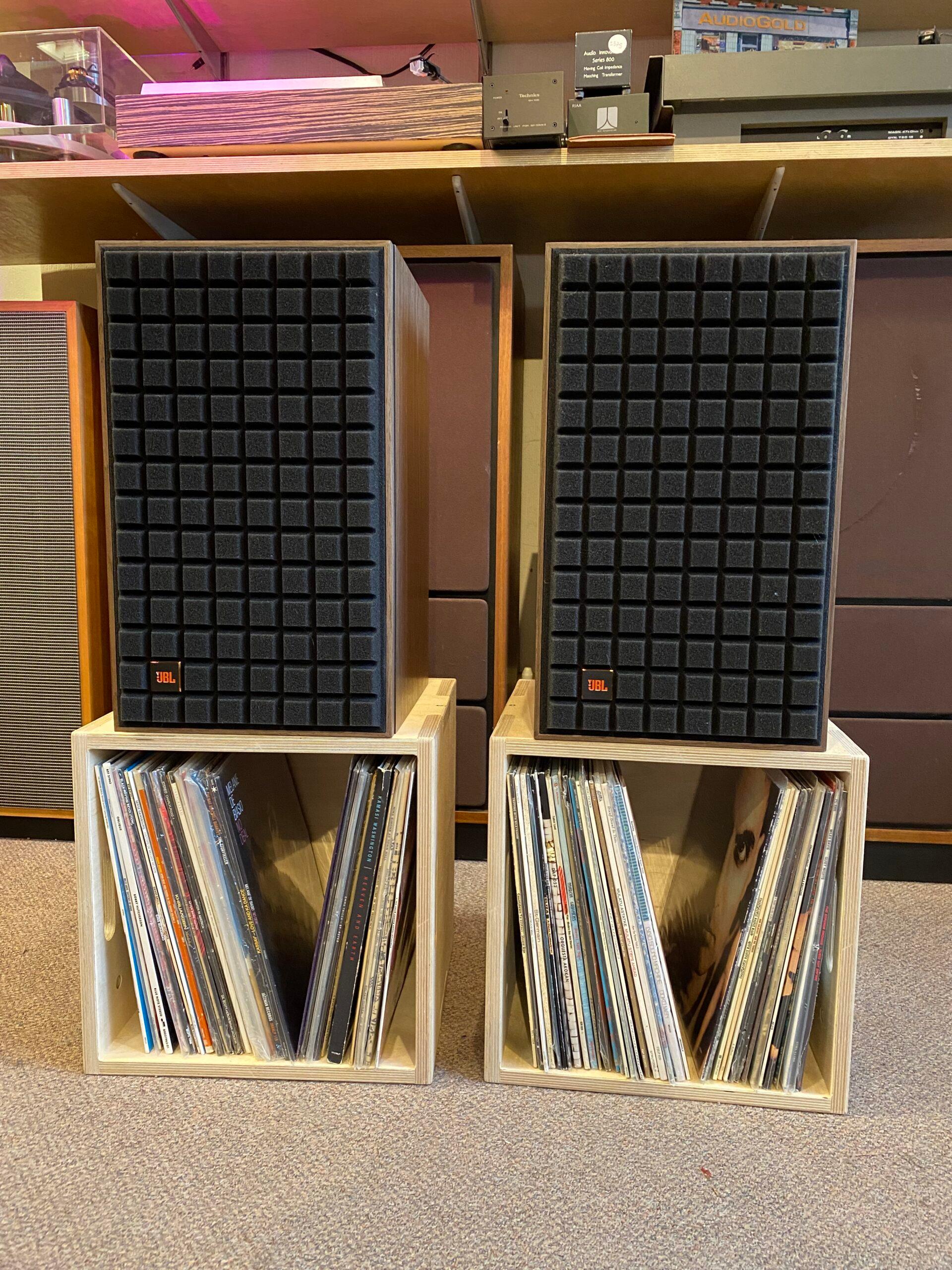 Two black speakers with grid-patterned fronts sit on top of upcycled wooden crates filled with vinyl records. The setup is on a carpeted floor, surrounded by other audio equipment and shelves in the background.