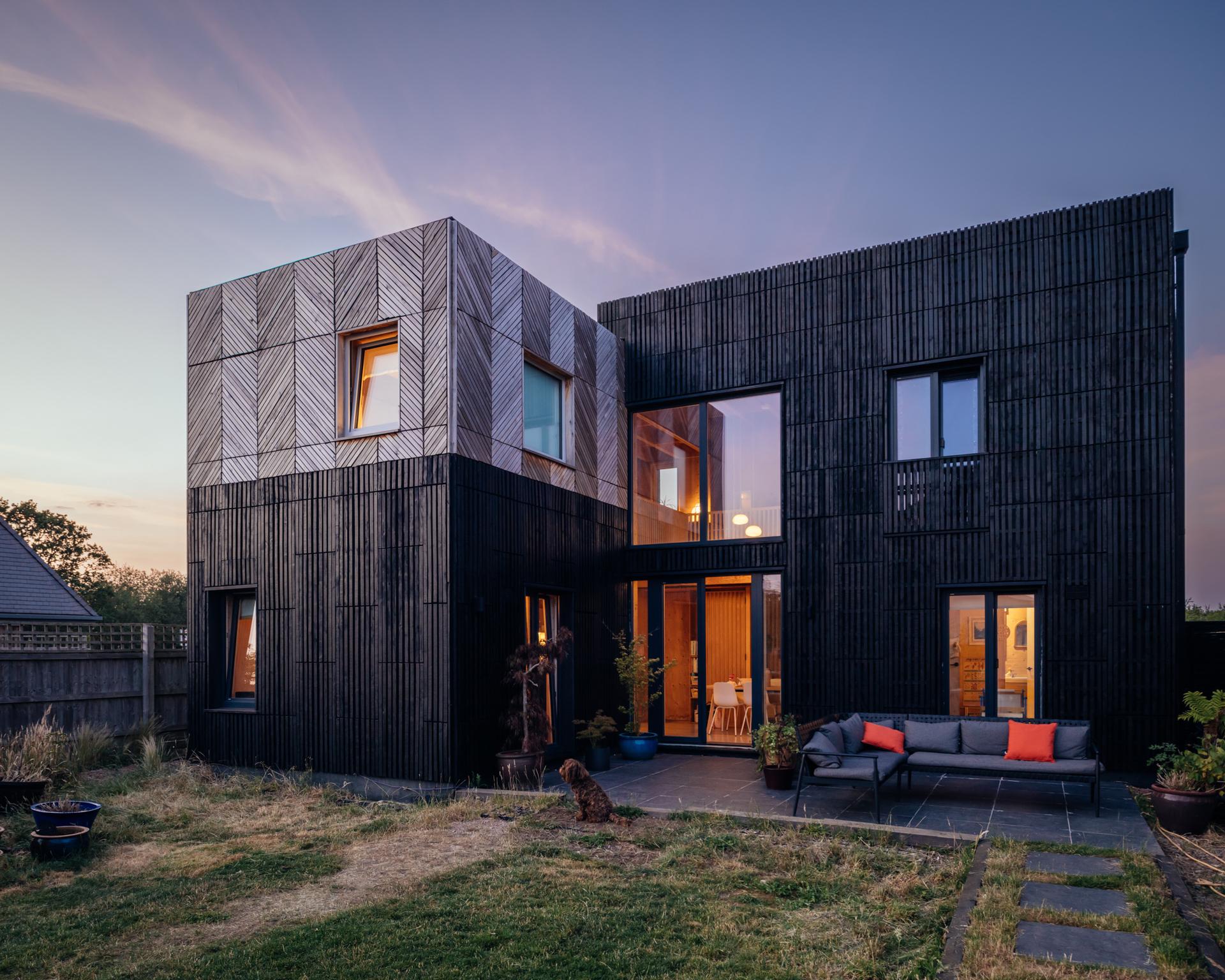 A modern two-storey home with a box extension featuring black slatted cladding and a patterned upper section, with a patio sectional sofa at dusk.