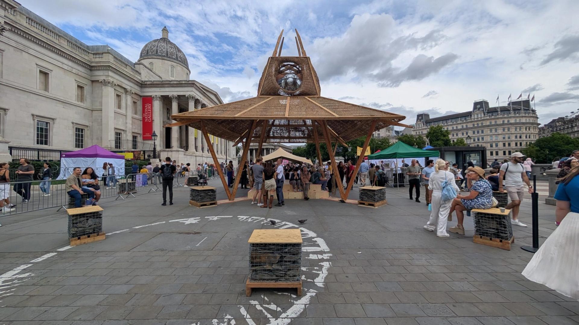 A wooden pavilion promoting sustainability with a mirrored sculpture at the top stands in Trafalgar Square, London. People walk and sit nearby; the National Gallery and colourful tents are visible in the background under a cloudy sky.