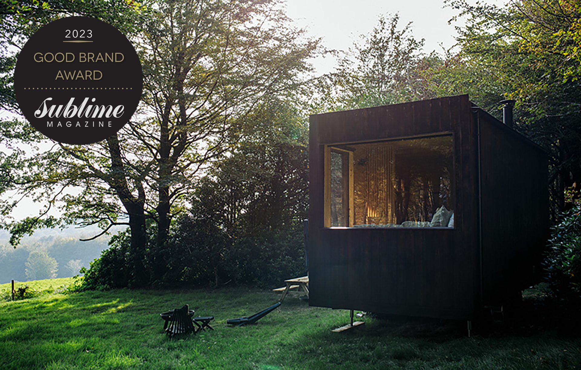 A modern, dark wooden cabin built with sustainably sourced materials sits in a grassy clearing surrounded by trees. A 2023 Good Brand Award, Sublime Magazine badge is displayed in the top left corner.