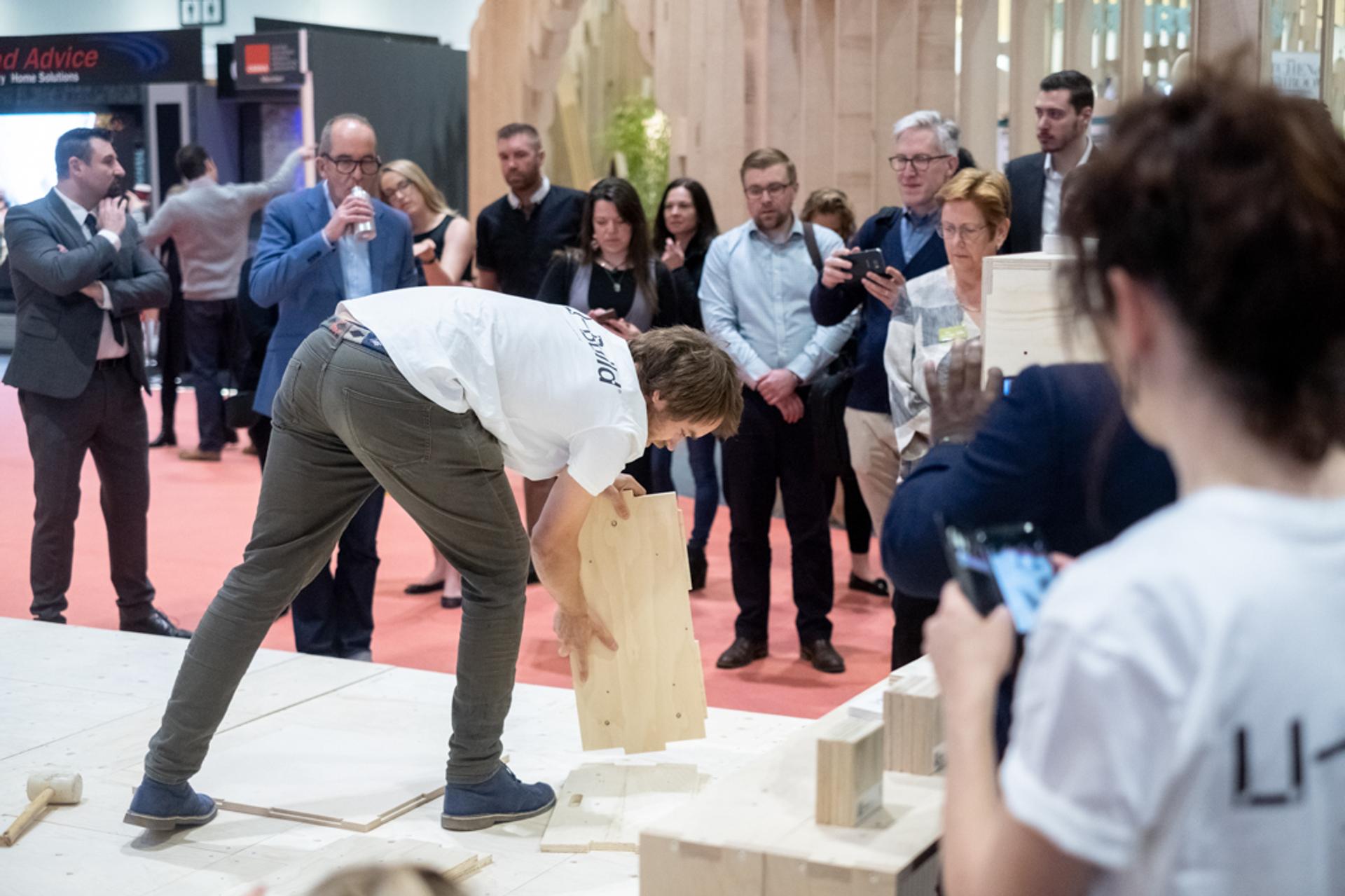 A person in a white shirt bends down to assemble modular wooden blocks on a platform, while a crowd of people stands around watching and taking photos at an indoor event or exhibition.