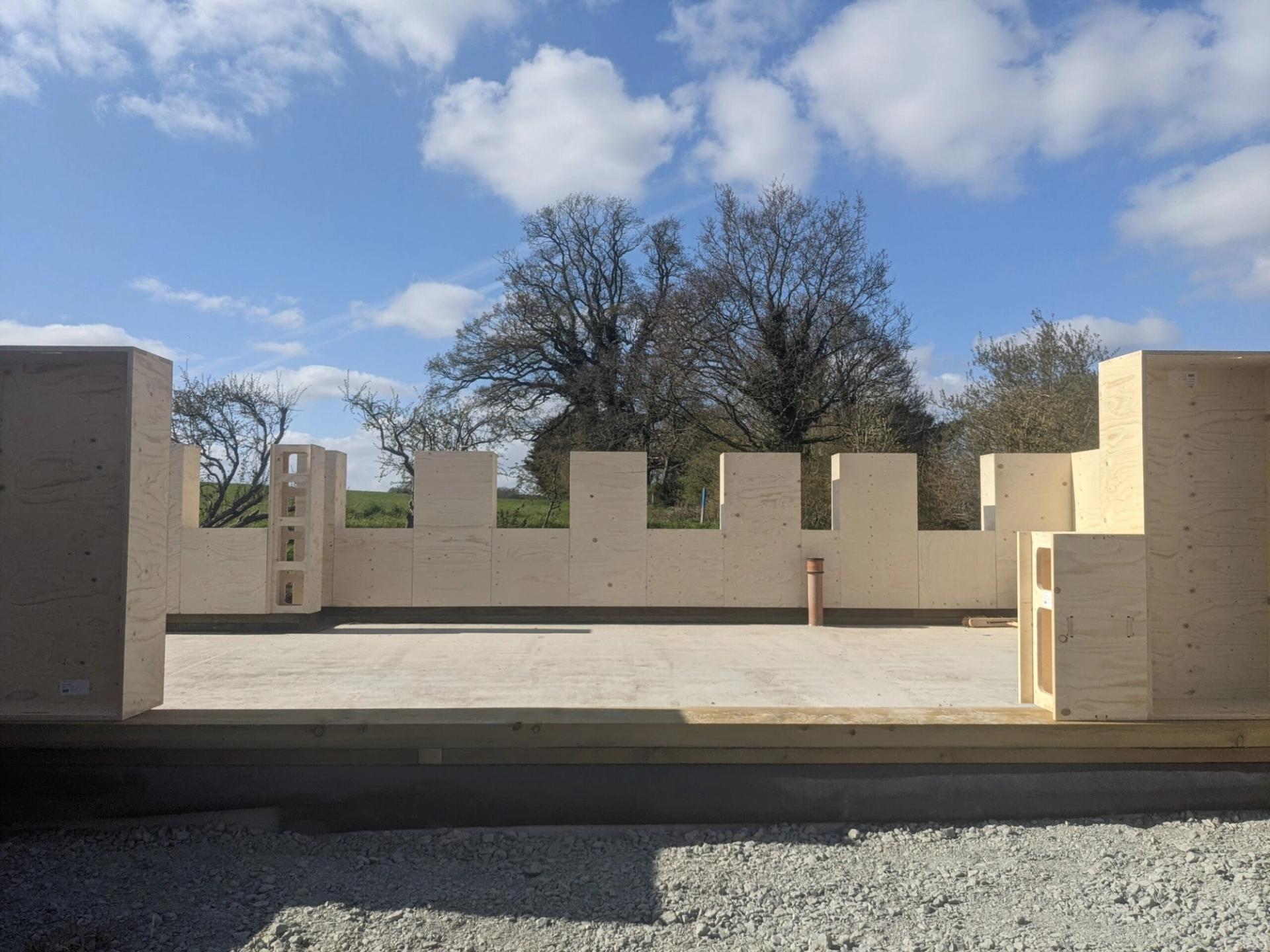 A building under construction with spruce timber wall panels partially installed, open window and door spaces visible, and trees and blue sky in the background, highlighting sustainable building practices.