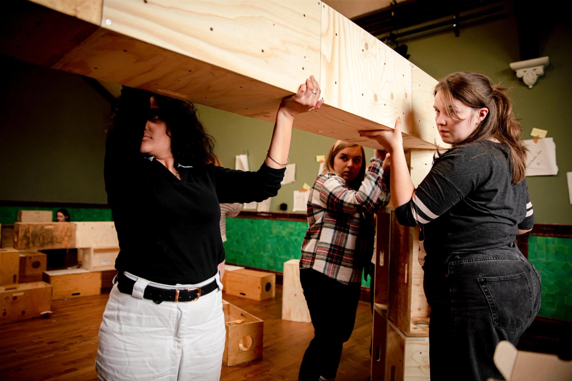 Three people work together to lift and assemble a large modular wooden structure indoors, surrounded by more spruce pieces and green tiled walls.