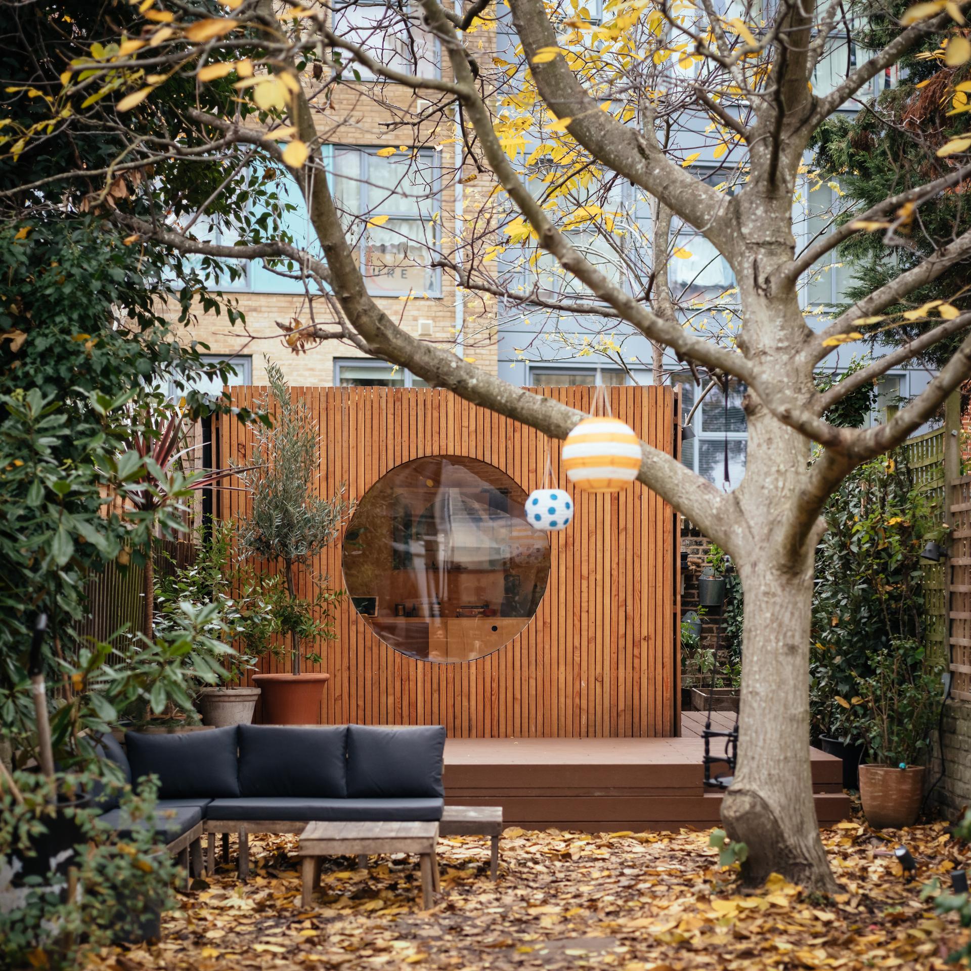 A cosy back garden with a sustainable timber construction garden room featuring a large circular window, surrounded by leafy trees. A black outdoor sofa and wooden bench sit on a patio covered with autumn leaves. Hanging ornaments decorate a spruce tree.