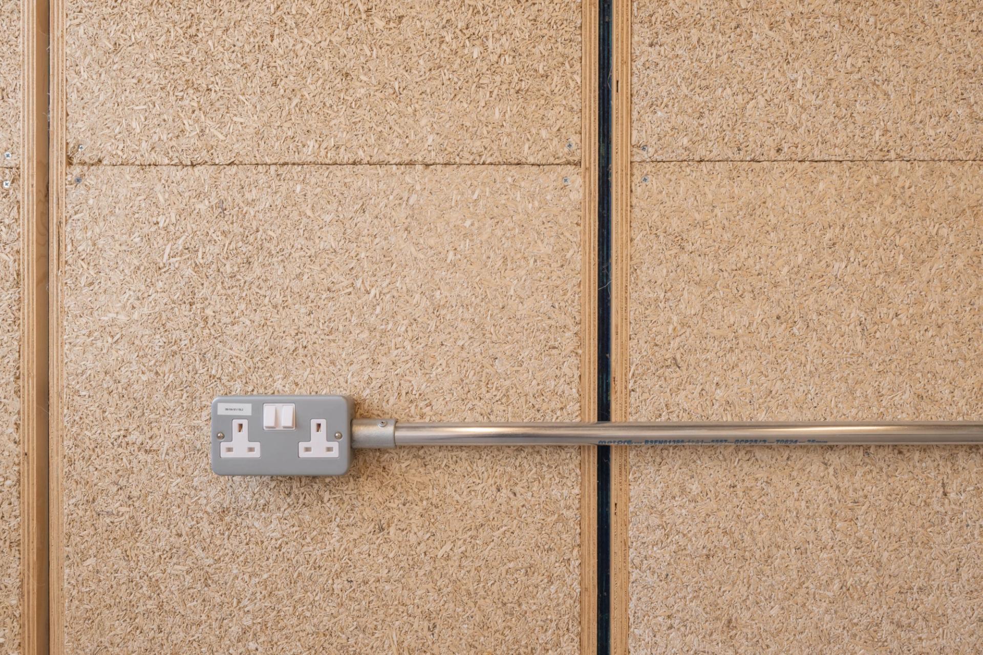 A double electrical socket with two switches is mounted on a light brown, textured wooden wall at Sustainable Ventures, Manchester, with a metal conduit running horizontally from the socket.