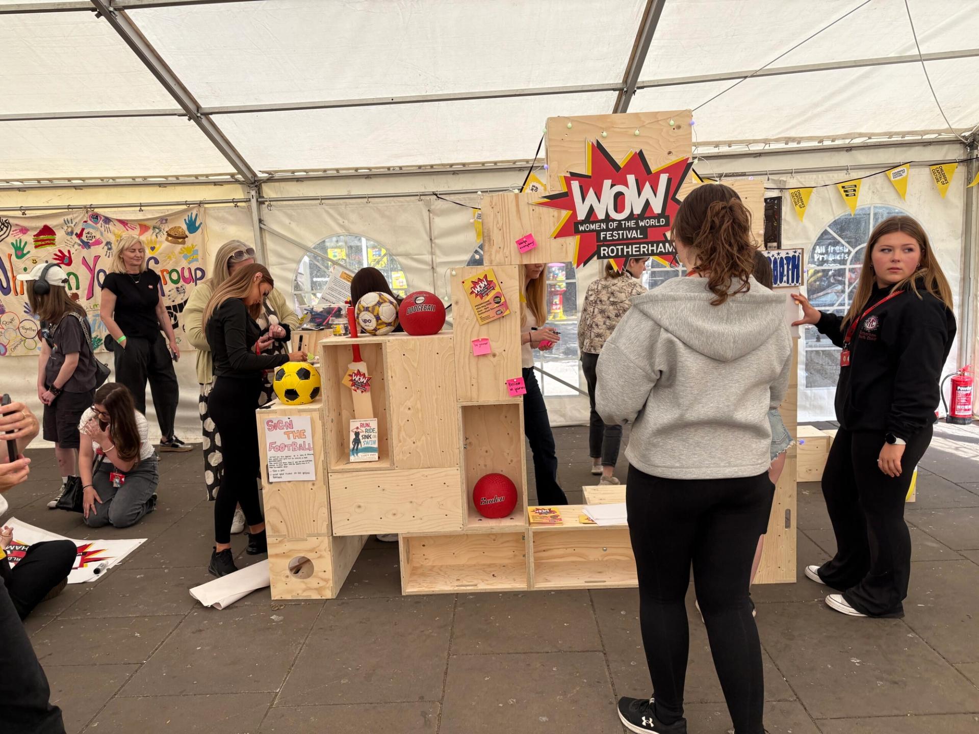 A group of people, mostly young women, gather around a timber construction display stand made of spruce with sports equipment and a WOW Women of the World Rotherham sign, inside a large white marquee at an event.
