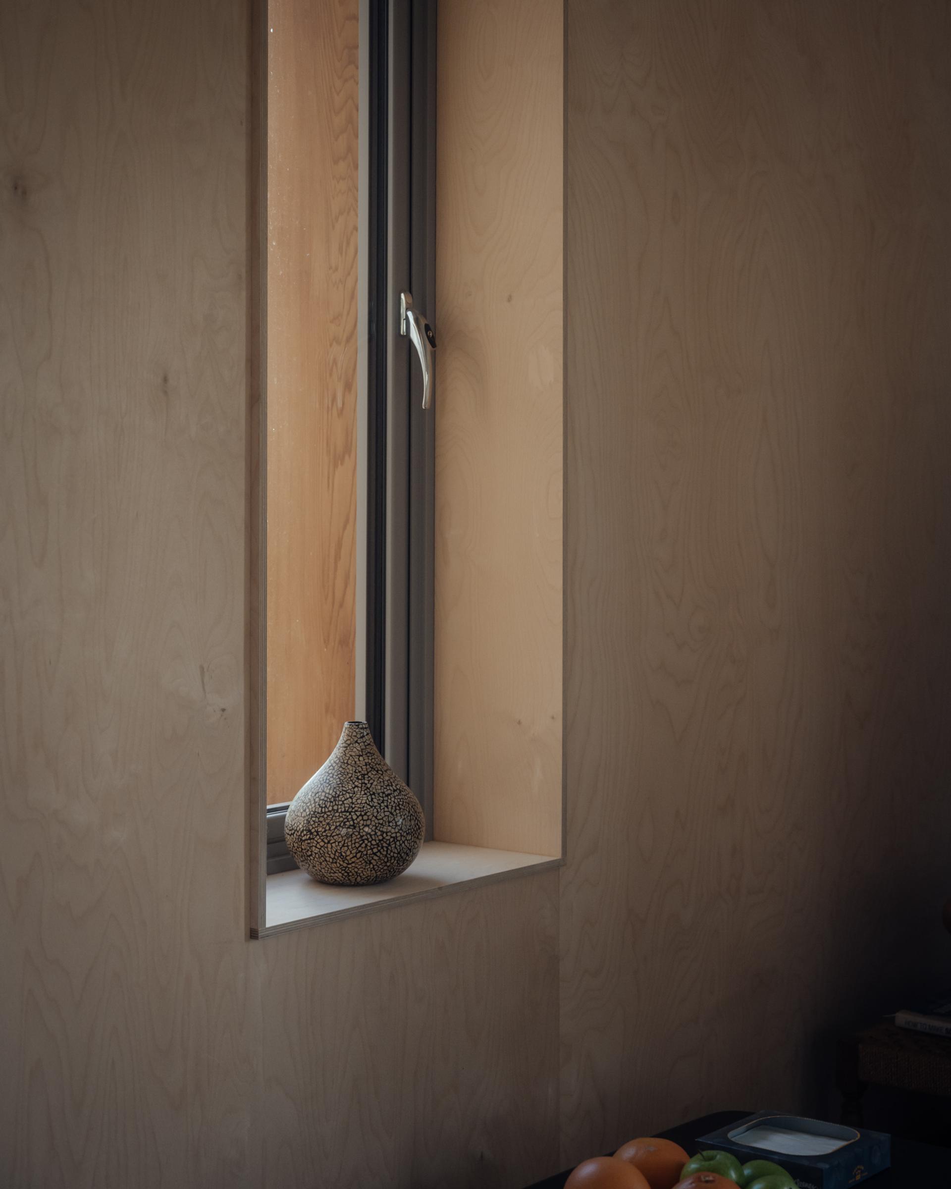 A patterned ceramic vase sits on a white windowsill in a minimalist room with light wood-panelled walls, highlighting plywood construction. A partially open window lets in soft daylight. Some fruit rests on a dark surface in the lower right corner.