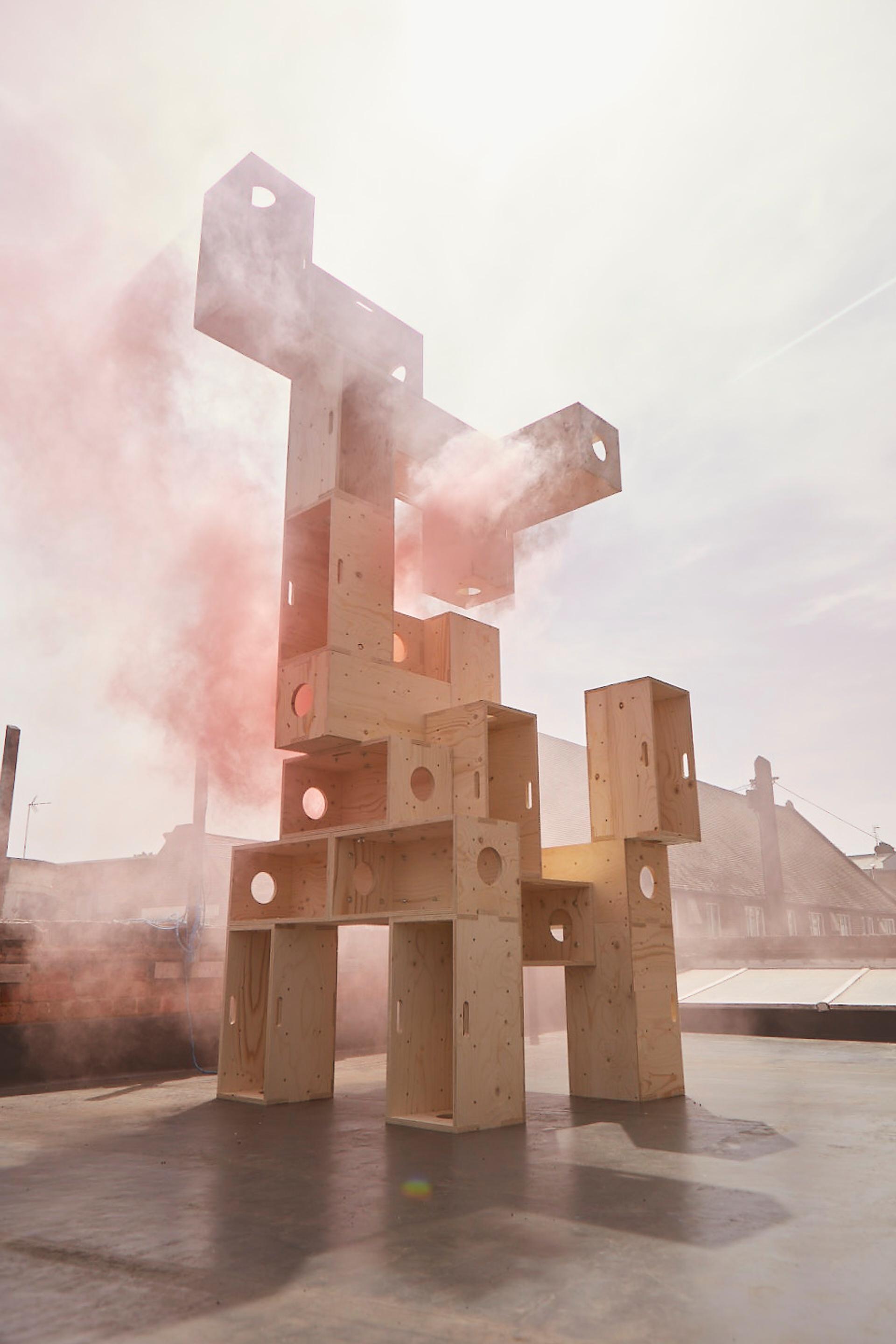 A tall, abstract wooden sculpture made of stacked modular rectangular blocks with circular cut-outs stands outdoors. Pink smoke surrounds the sustainable timber construction as sunlight shines through, casting shadows on the ground.