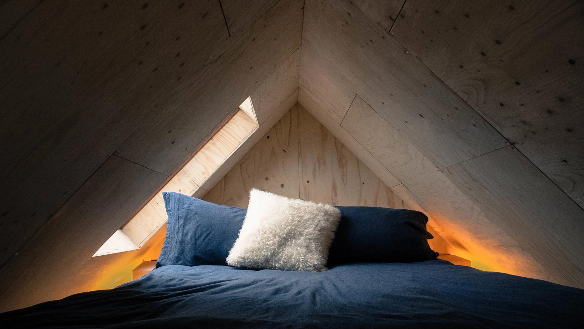 A cosy bed with dark blue sheets and a white fluffy pillow is positioned under a wooden A-frame ceiling made from sustainably sourced materials, with small triangular windows letting in soft light.