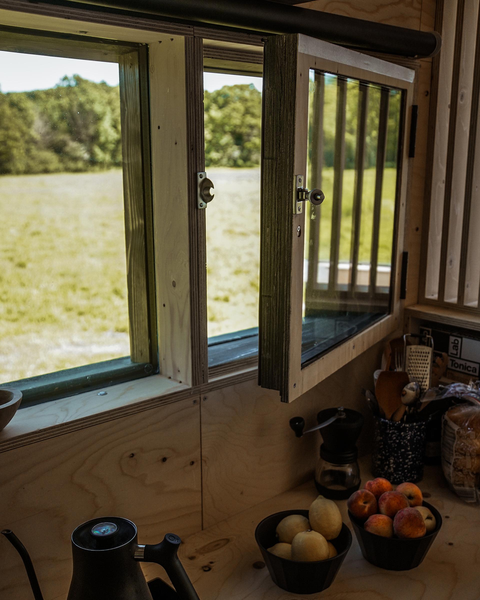 A kitchen worktop with fruit bowls, utensils, and a kettle sits beneath an open window, offering a sunny view of grassy fields and green trees—a perfect space to embrace sustainability in everyday living.