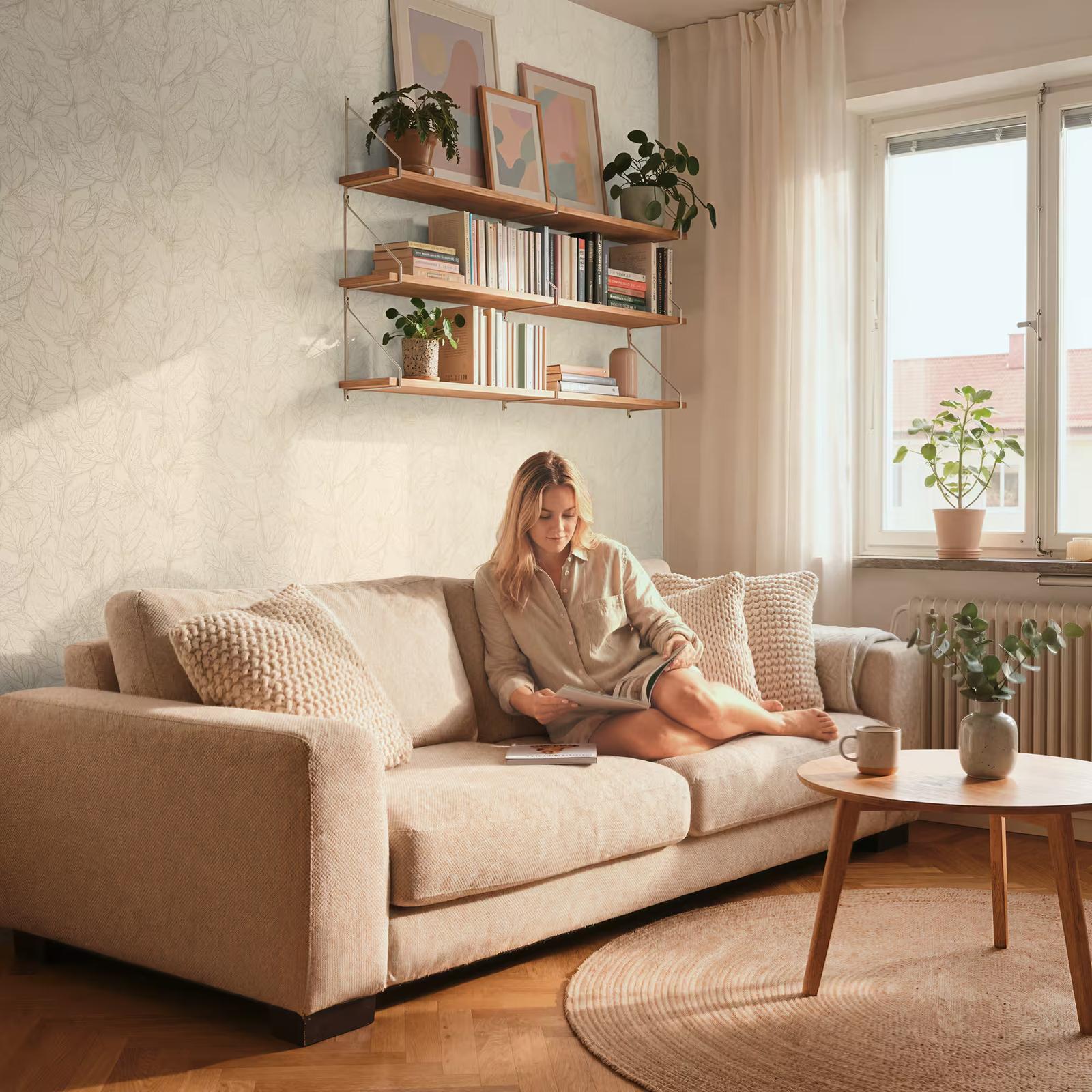 A woman reading on a beige sofa against pale line-drawn botanical wallpaper, below shelves of books and plants