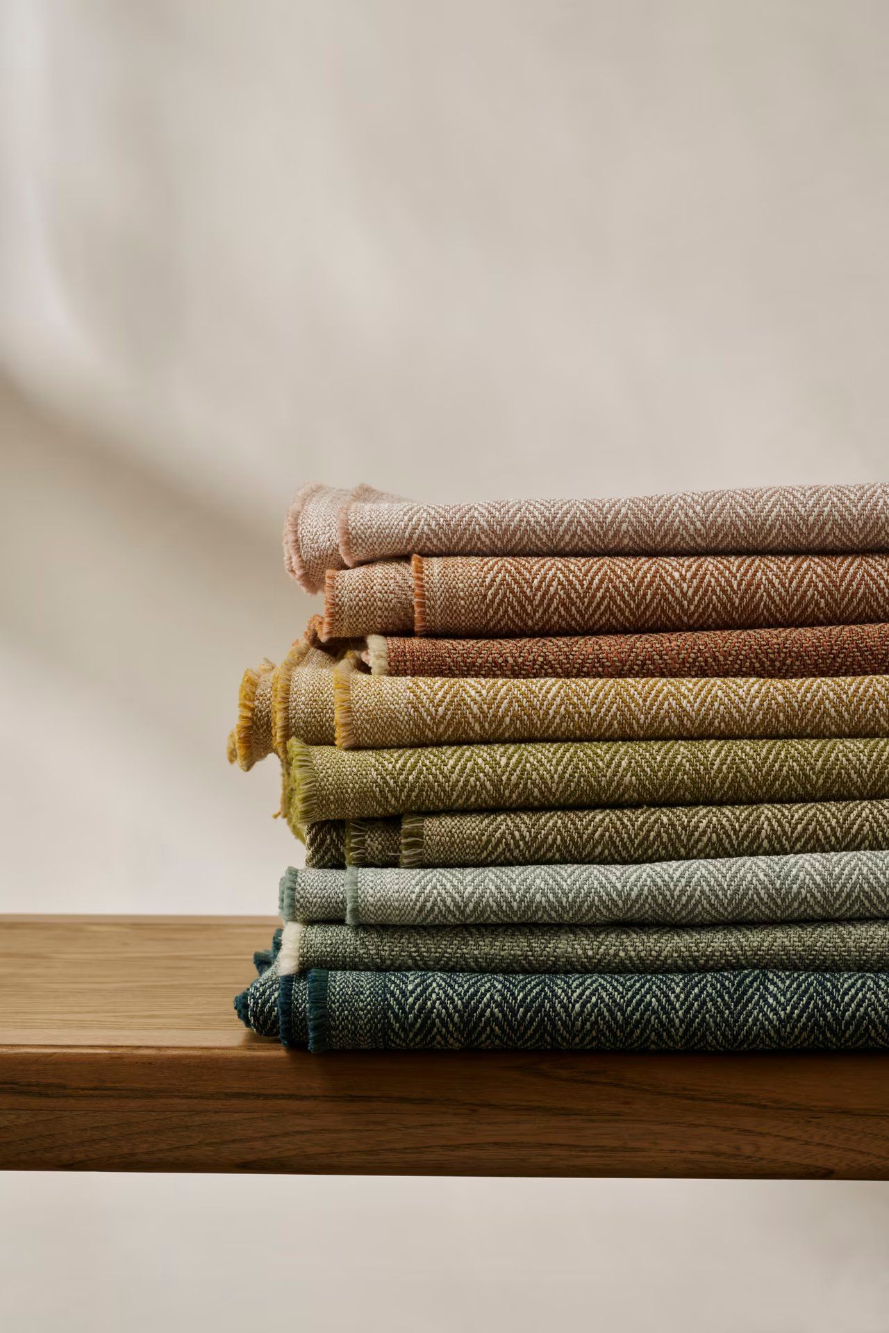 Folded herringbone upholstery fabrics in pink, ochre, olive and teal stacked on an oak shelf