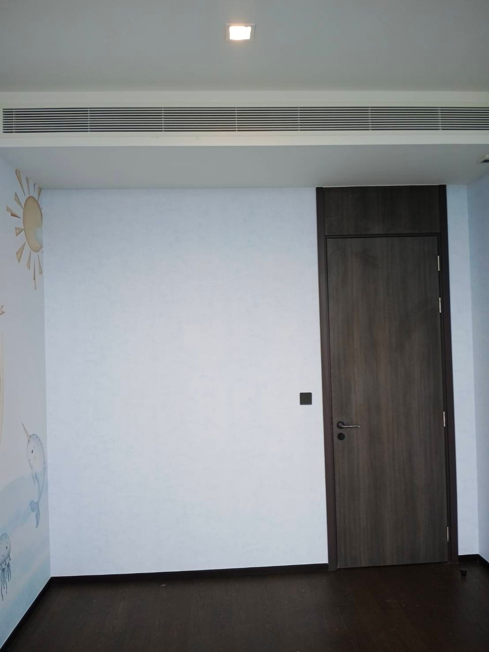 A full wall of Sky Wash plain wallpaper beside the nursery door, the mural's sun and narwhal just visible at the left-hand corner
