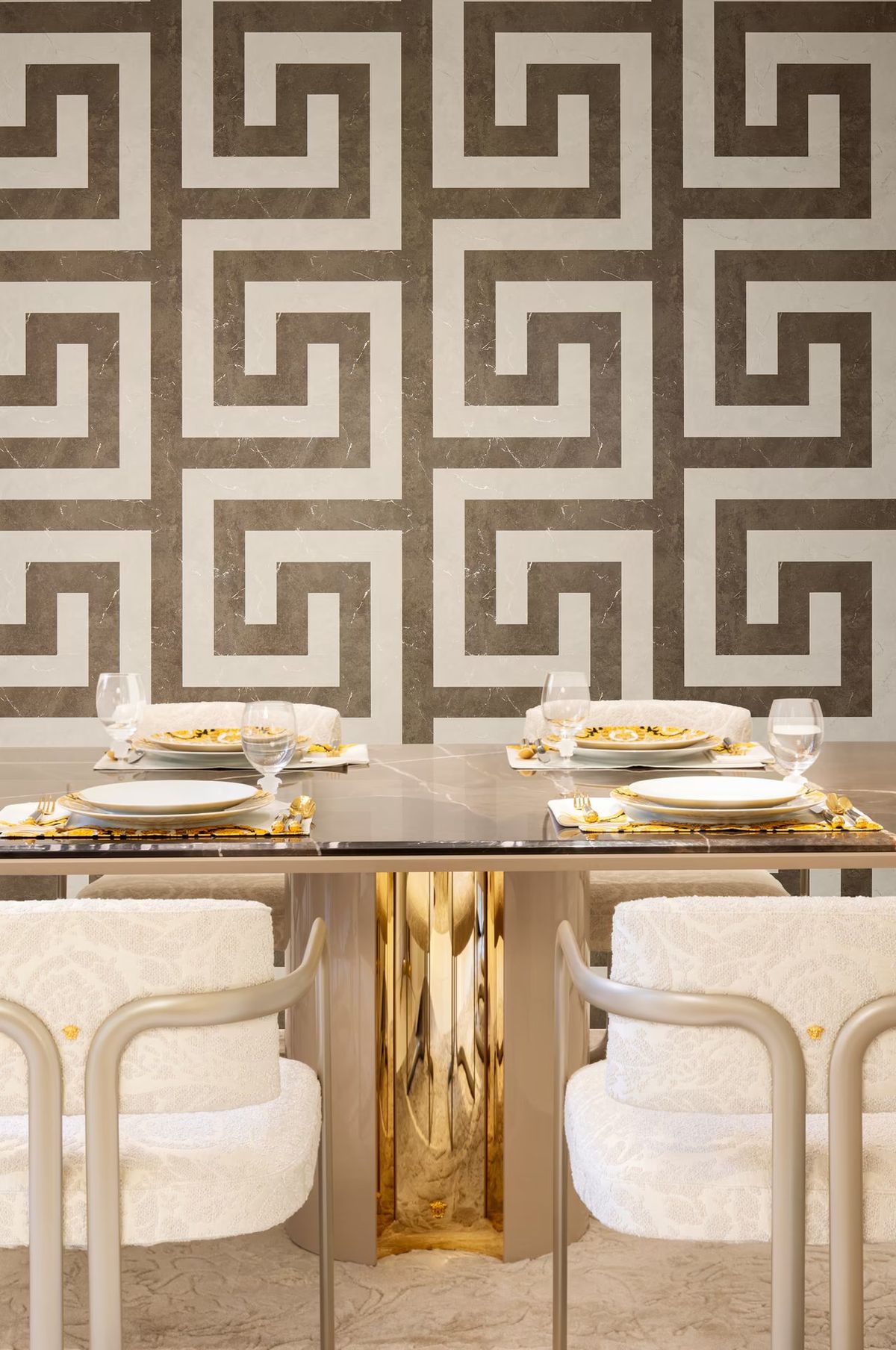 Grey and cream Greek key wallpaper behind a dining table laid with gold-rimmed plates and bouclé chairs