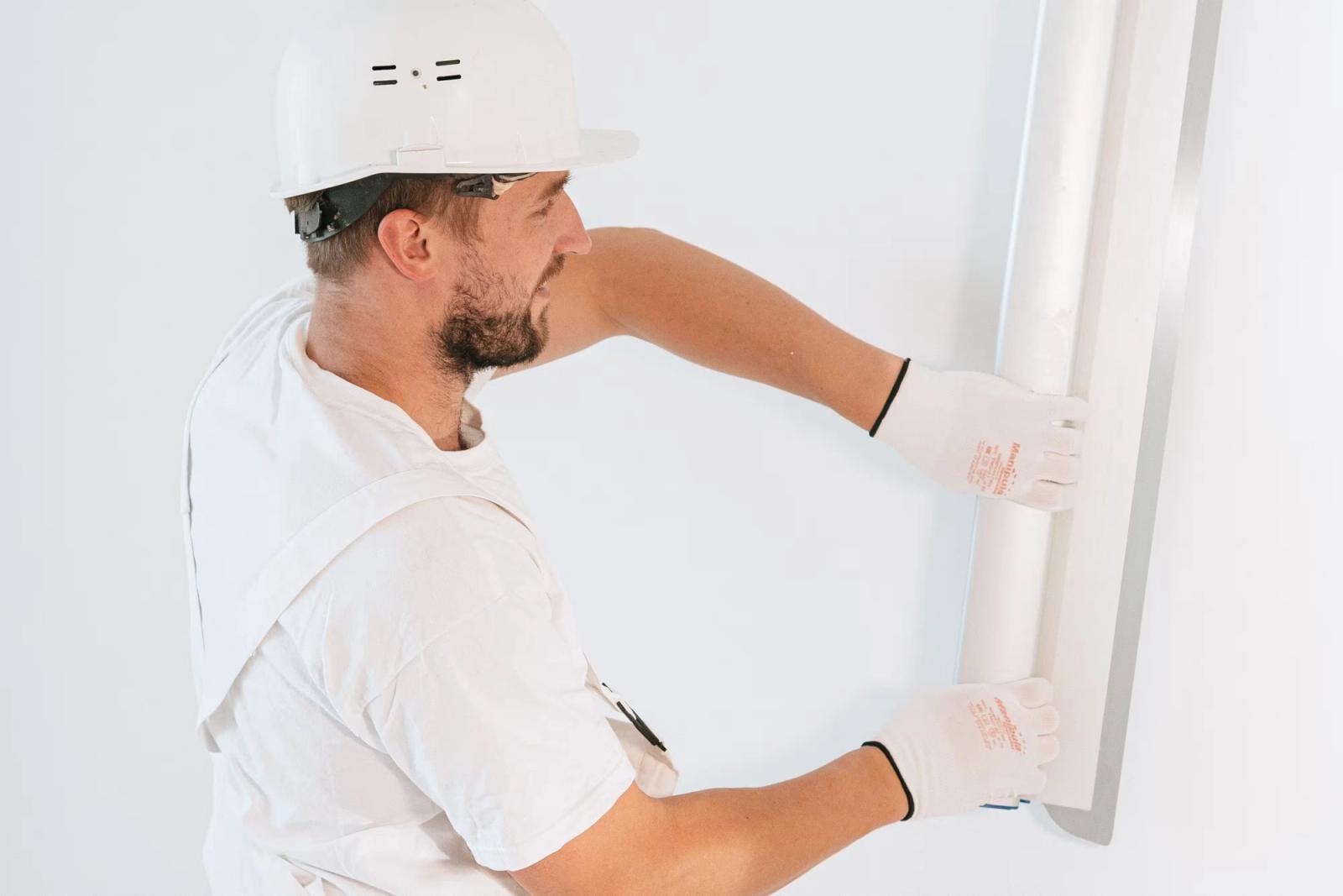 An installer in white overalls and a hard hat holding a rolled length of wallpaper against a plain white wall