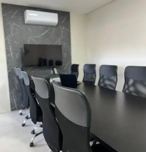 Sala de reunião com mesa longa preta, cadeiras ergonômicas e tela na parede para apresentações