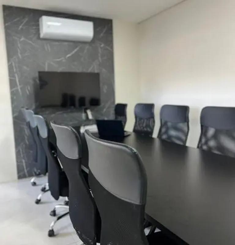 Sala de reunião com mesa longa preta, cadeiras ergonômicas e tela na parede para apresentações