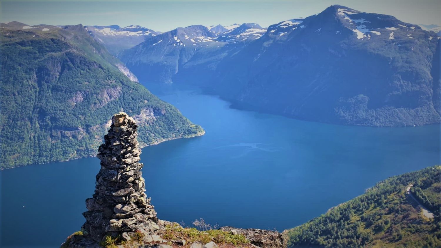 Panoramablick auf den Storfjorden in Sunnmøre, Norwegen, mit tiefblauem Fjordwasser, hoch aufragenden Bergen und einem traditionellen Steinhaufen am Aussichtspunkt.