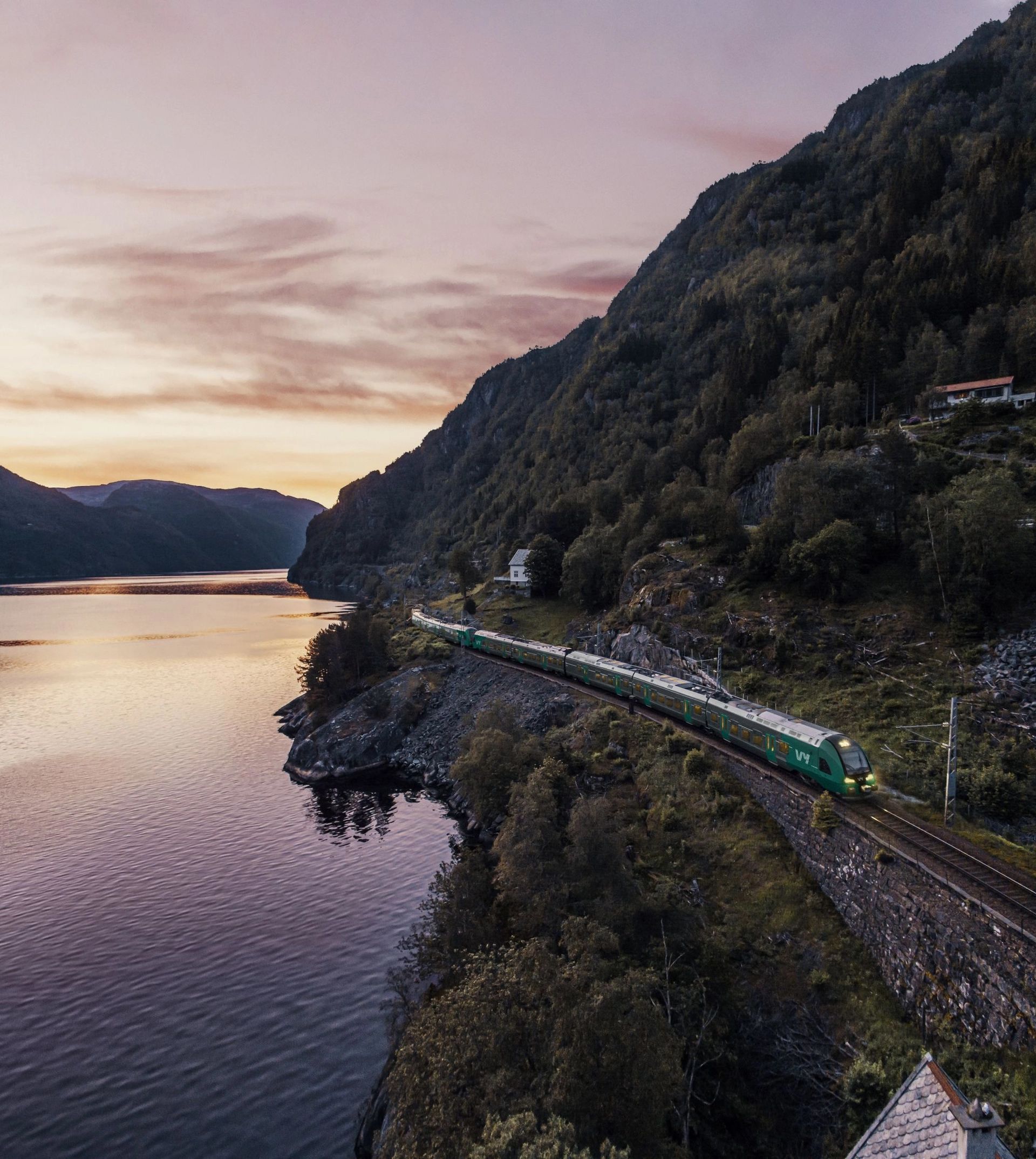 Zug der Bergenbahn, der bei Sonnenuntergang in Norwegen an einem Fjord entlangfährt.