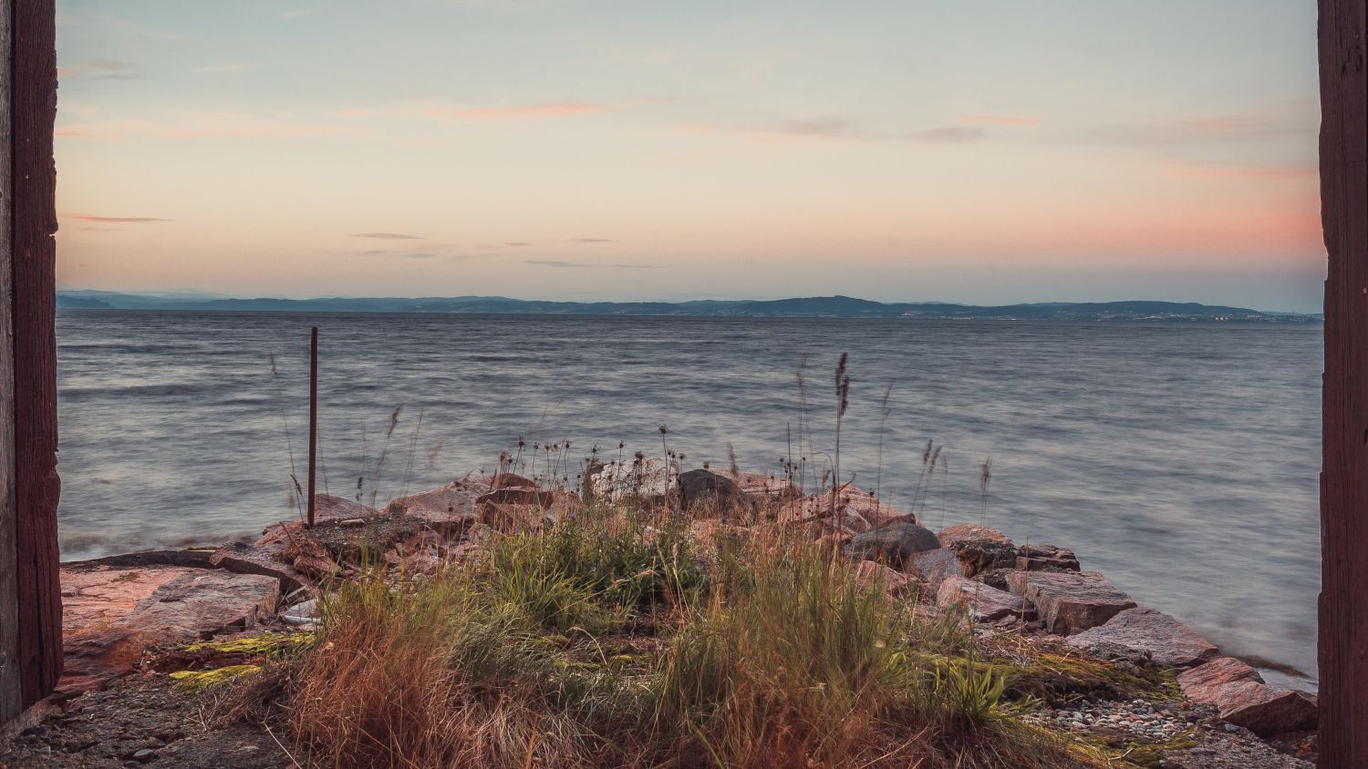 Friedlicher Sonnenuntergang über dem Trondheimsfjord mit felsiger Küste, sanften Wellen und einem pastellfarbenen Abendhimmel in Zentralnorwegen.