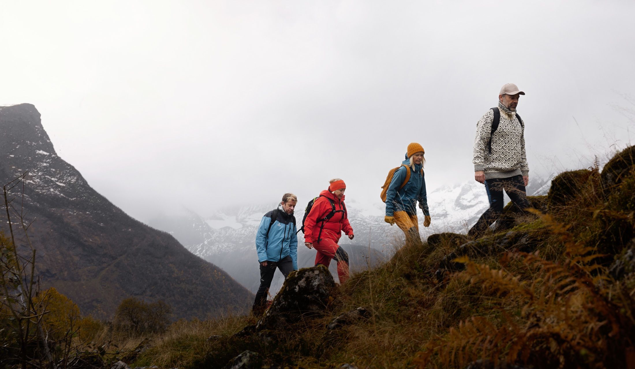 Gruppe von vier Wanderern in bunten Jacken, die auf einem Bergpfad wandern, mit nebelverhangenen, schneebedeckten Gipfeln im Hintergrund bei einer Bergwanderung auf einem Bauernhof im Hjørundfjord in Norwegen
