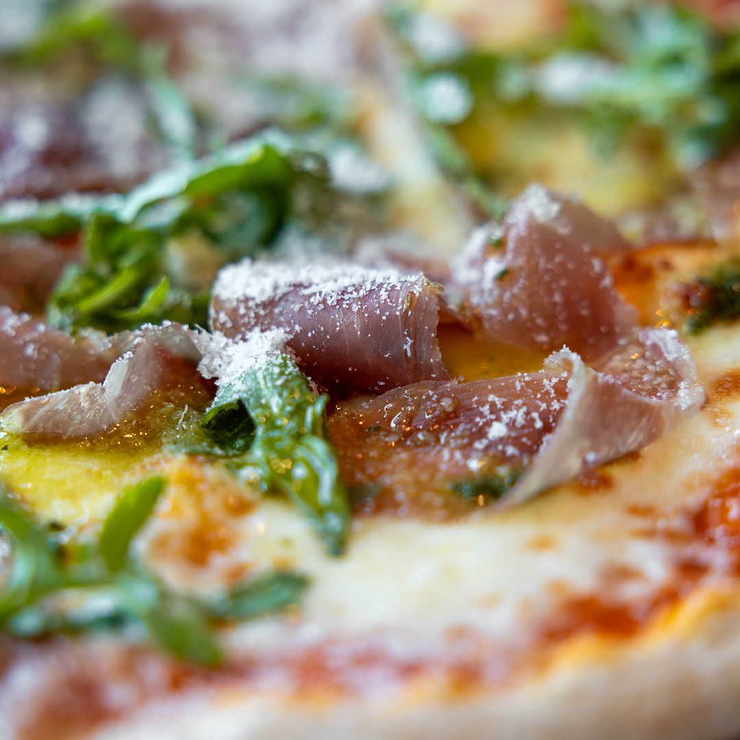 Närbild på en pizza toppad med lufttorkad skinka, ruccola och riven ost på Havly Cafe