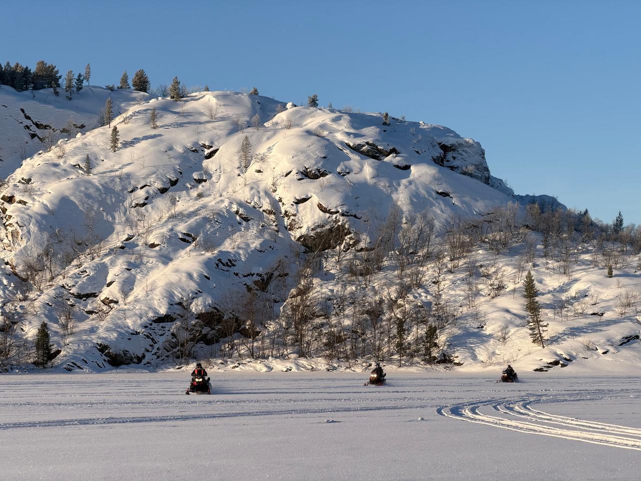 Three snowmobiles crossing a frozen lake below a snow-covered hillside on a snowmobile safari in Kirkenes, Norway