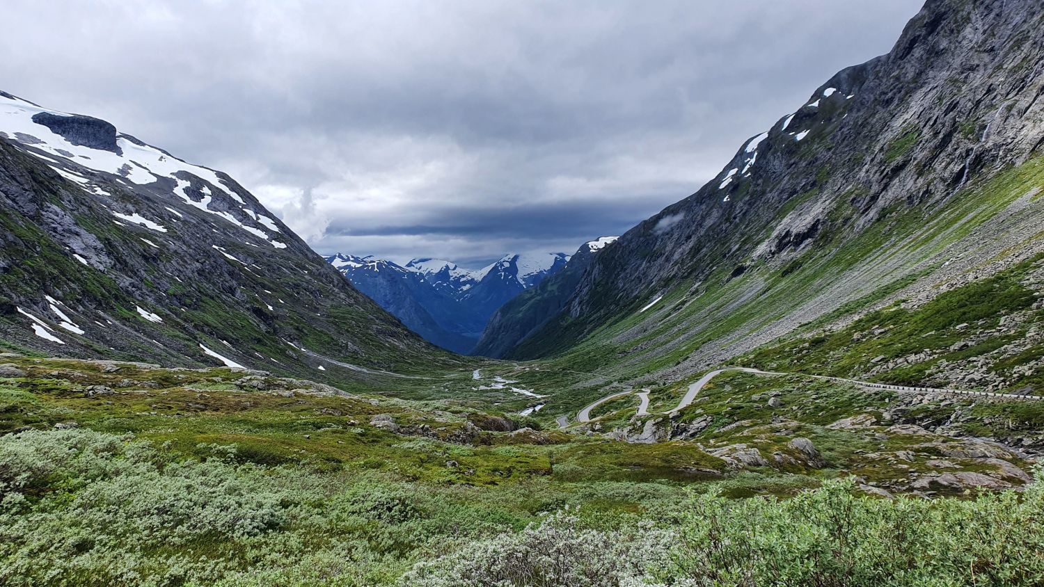 Malerisches Hochtal mit einer kurvenreichen Straße, umgeben von imposanten Gipfeln und Schneefeldern in den norwegischen Highlands