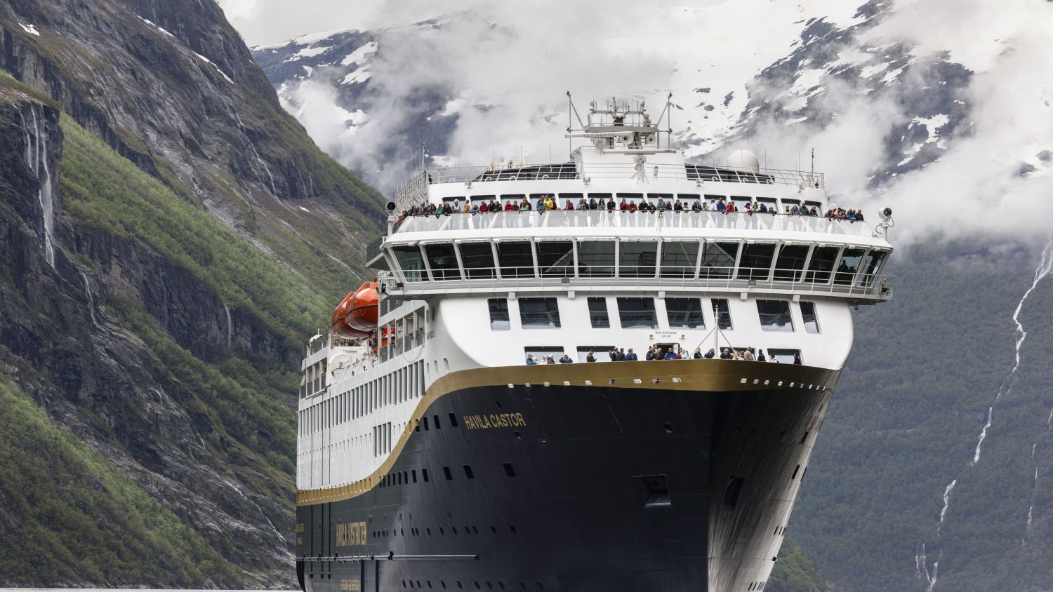 Havila Castor seiler gjennom Geirangerfjorden, omgitt av ruvende klipper, fossefall og passasjerer som nyter den panoramiske utsikten fra dekk