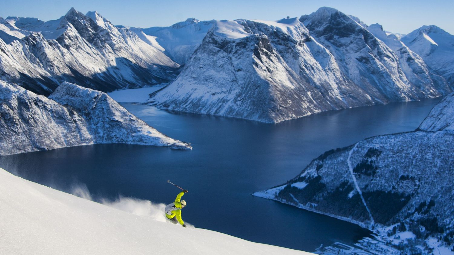 Skifahrer, der einen verschneiten Berg oberhalb des Hjørundfjords hinabfährt, umgeben von dramatischen schneebedeckten Gipfeln und dem tiefblauen Fjord in Westnorwegen.