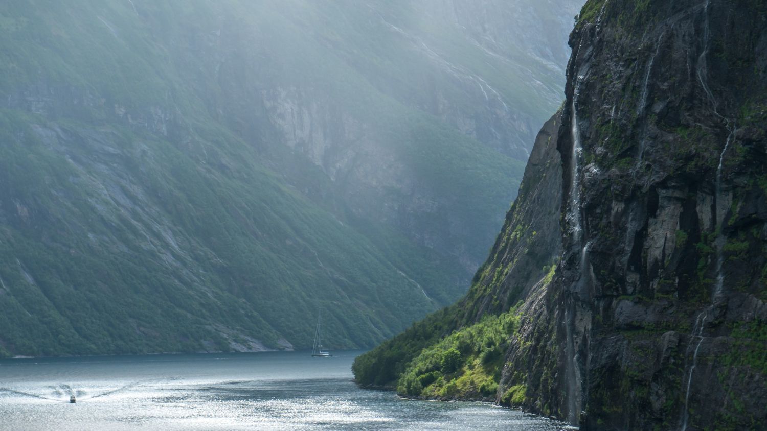 Sollys som filtrerer ned i Geirangerfjorden og lyser opp ruvende klipper, brusende fossefall og rolig fjordvann omgitt av frodige grønne fjell