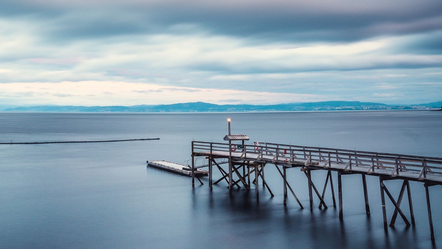 Hölzerner Steg, der in das ruhige Wasser des Trondheimsfjords in Trøndelag, Norwegen, hinausragt, unter einem dramatisch bewölkten Himmel während der Blauen Stunde.