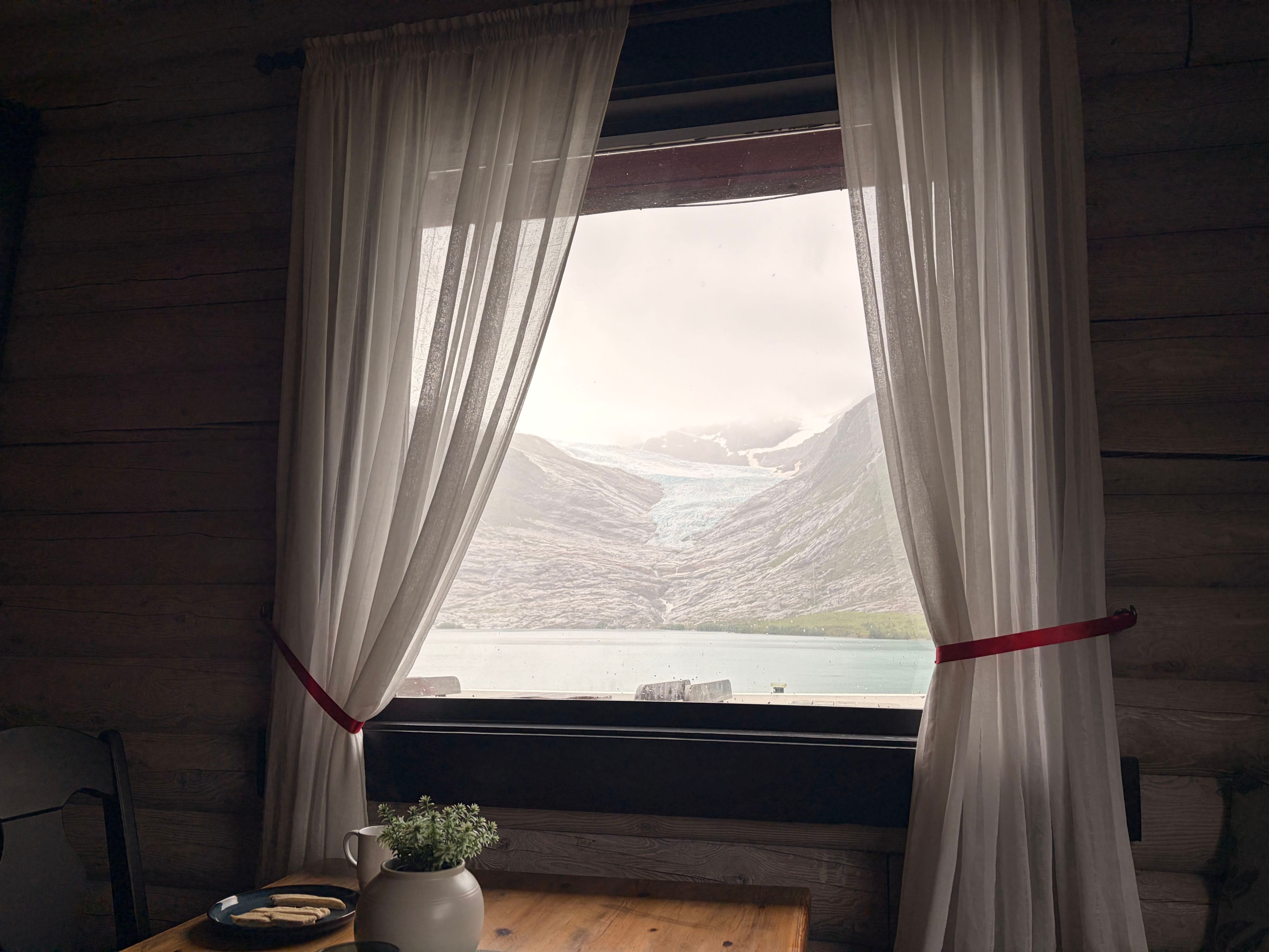 Der Engabreen-Gletscher und sein Schmelzwassersee, gesehen durch das von Vorhängen eingerahmte Fenster des Blockhüttencafés am Svartisen, mit Kaffee und Waffeln auf dem Tisch