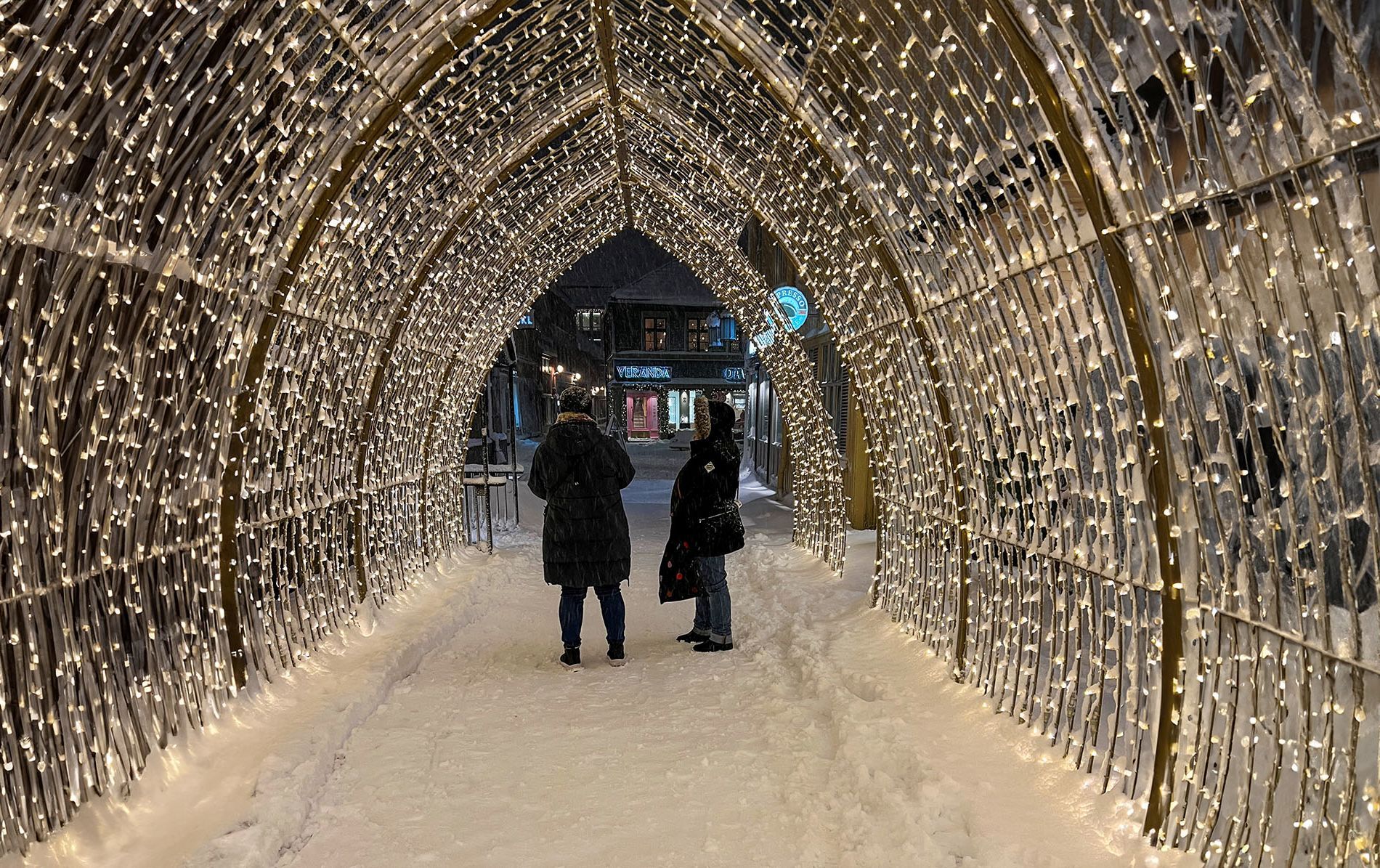 Two people standing in a glowing Christmas light tunnel on a snowy street at night in Trondheim, Norway