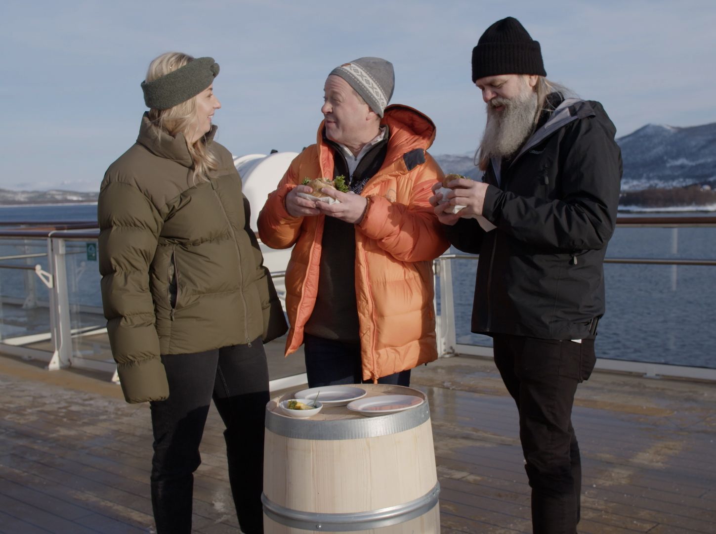 Tre gjester i vinterjakker smaker på mat fra små tallerkener på en tønne på skipsdekket