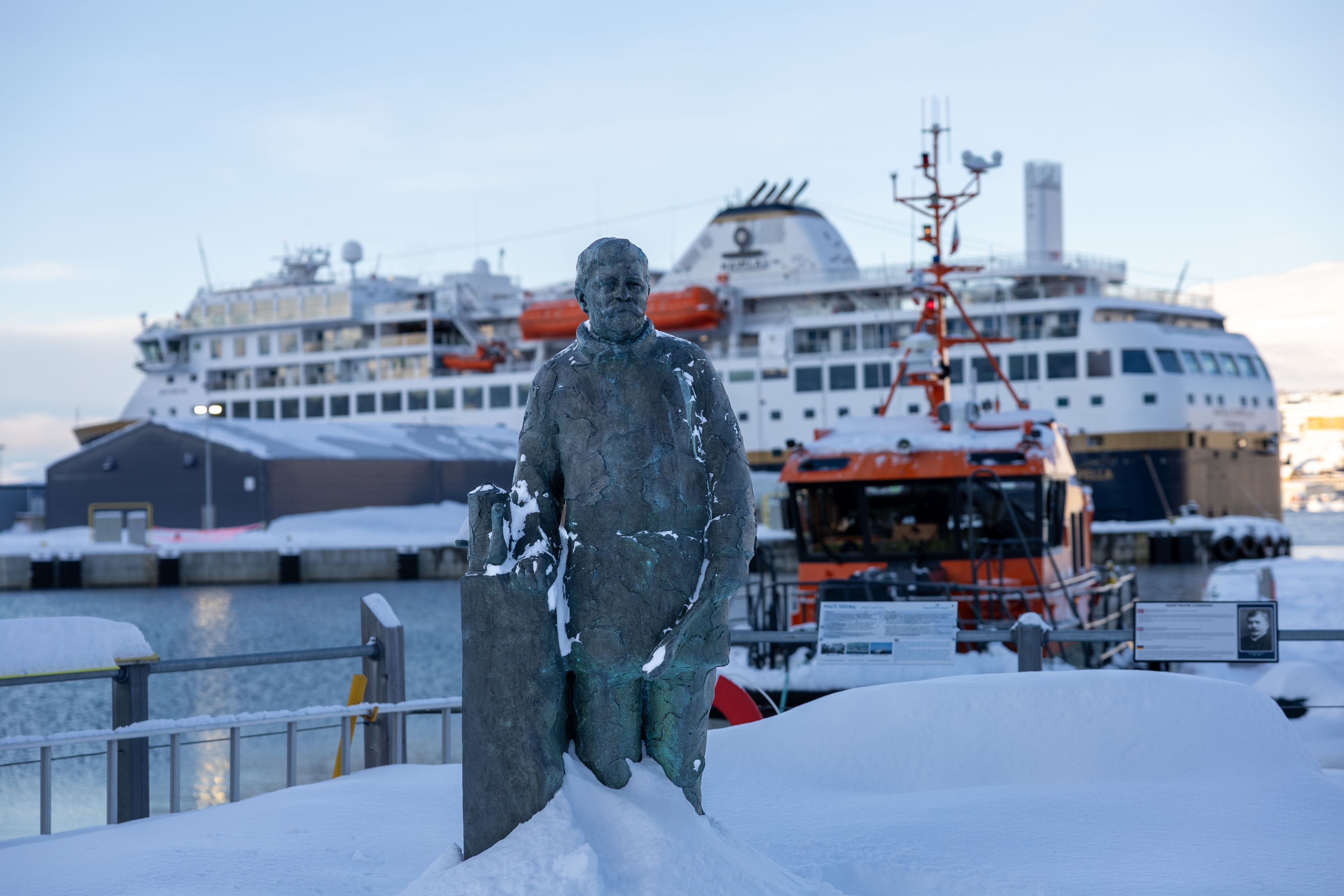 Snødekt bronsestatue i Hammerfest havn med et cruiseskip og en losbåt bak på en vinterbyvandring, Norge