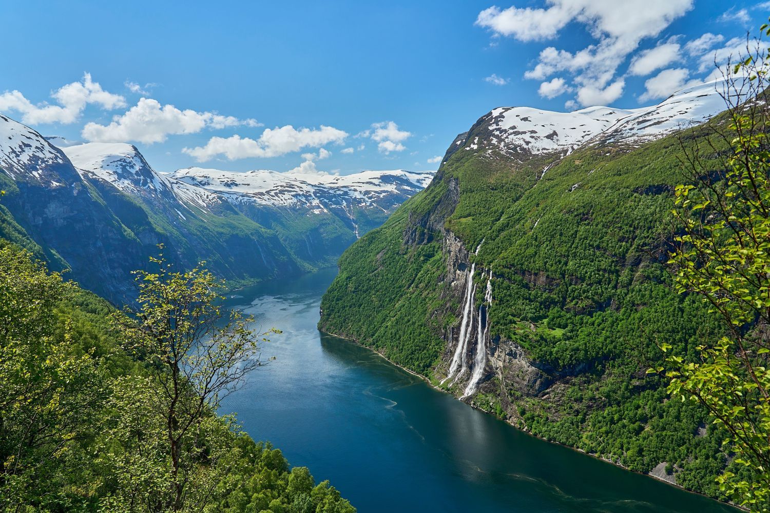 Cruiseskip som seiler gjennom Geirangerfjorden om sommeren, omgitt av smaragdgrønt vann, ruvende klipper, frodige grønne fjell og fossefall som stuper nedover fjellsidene