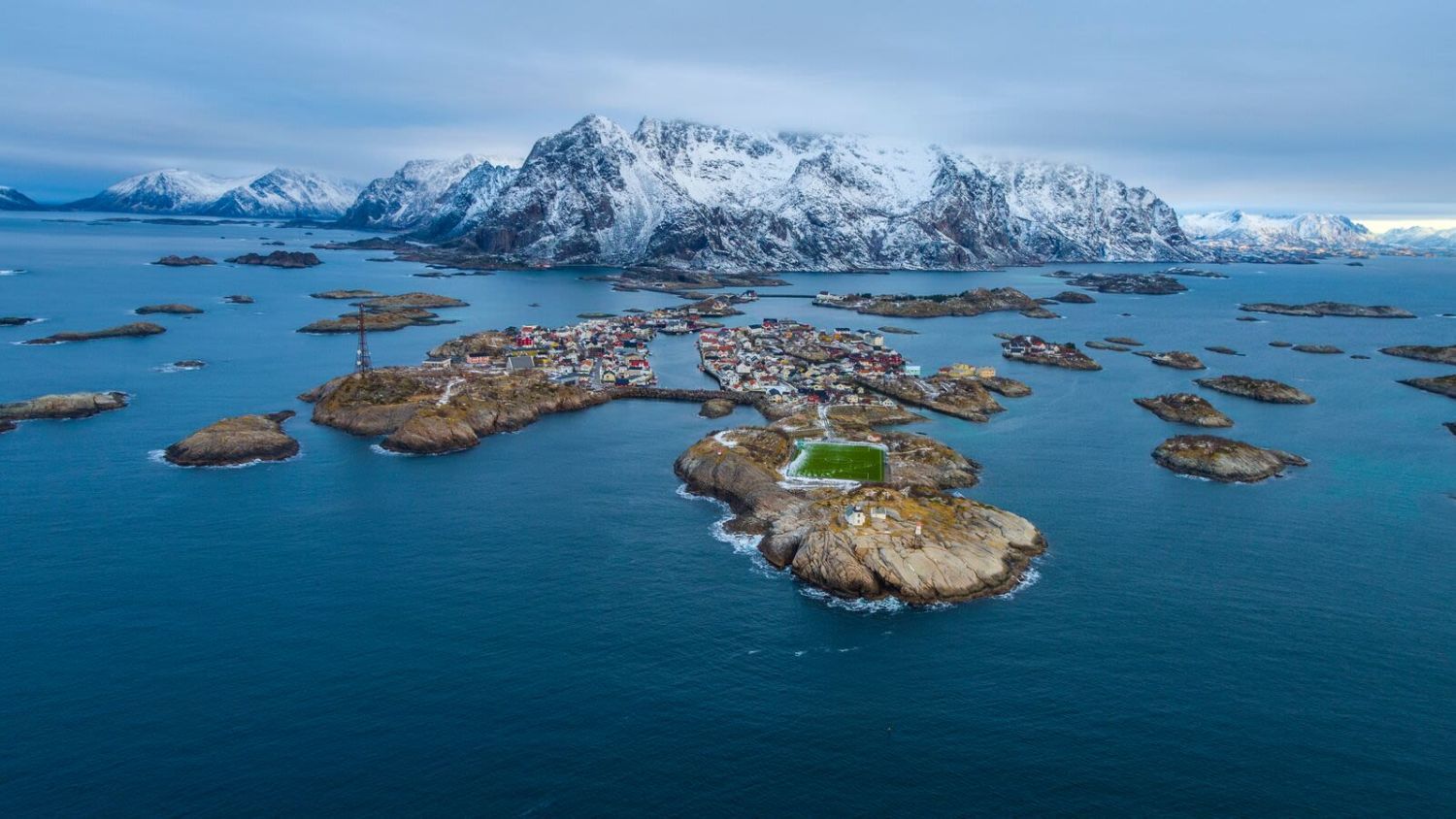 Luftaufnahme von Henningsvær auf den Lofoten, umgeben von zerklüfteten Schären, dem Norwegischen Meer und schneebedeckten Bergen
