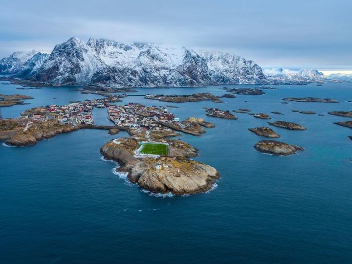 Luftaufnahme von Henningsvær auf den Lofoten, umgeben von zerklüfteten Schären, dem Norwegischen Meer und schneebedeckten Bergen