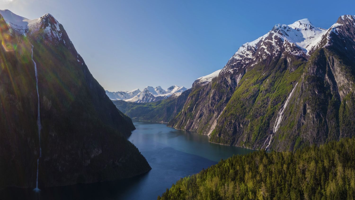 Panoramablick auf den Storfjord in Sunnmøre, Norwegen, mit schneebedeckten Bergen, rauschenden Wasserfällen, üppigen Wäldern und tiefblauem Fjordwasser.